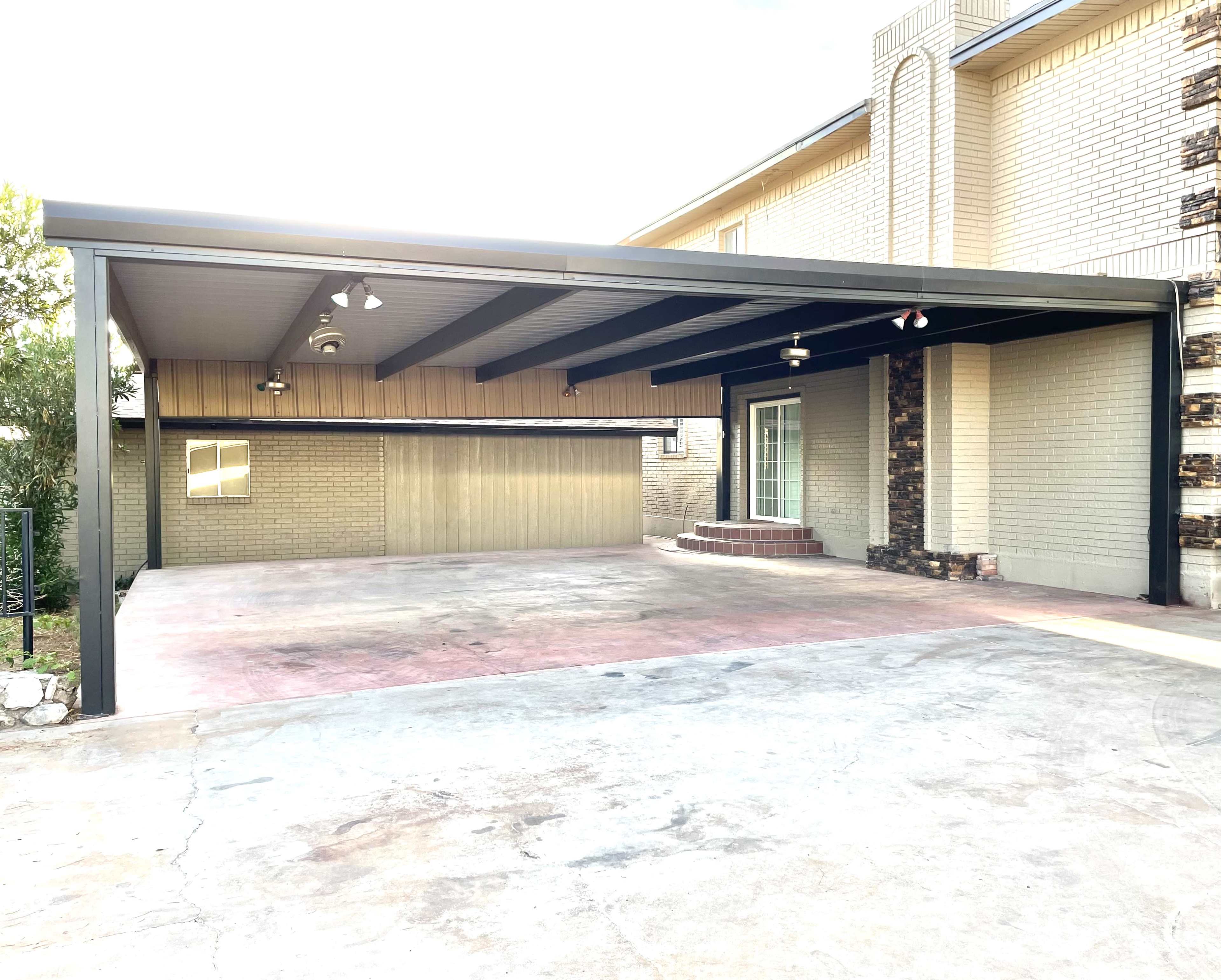 A large, covered carport attached to a brick house, with a concrete driveway leading up to it.