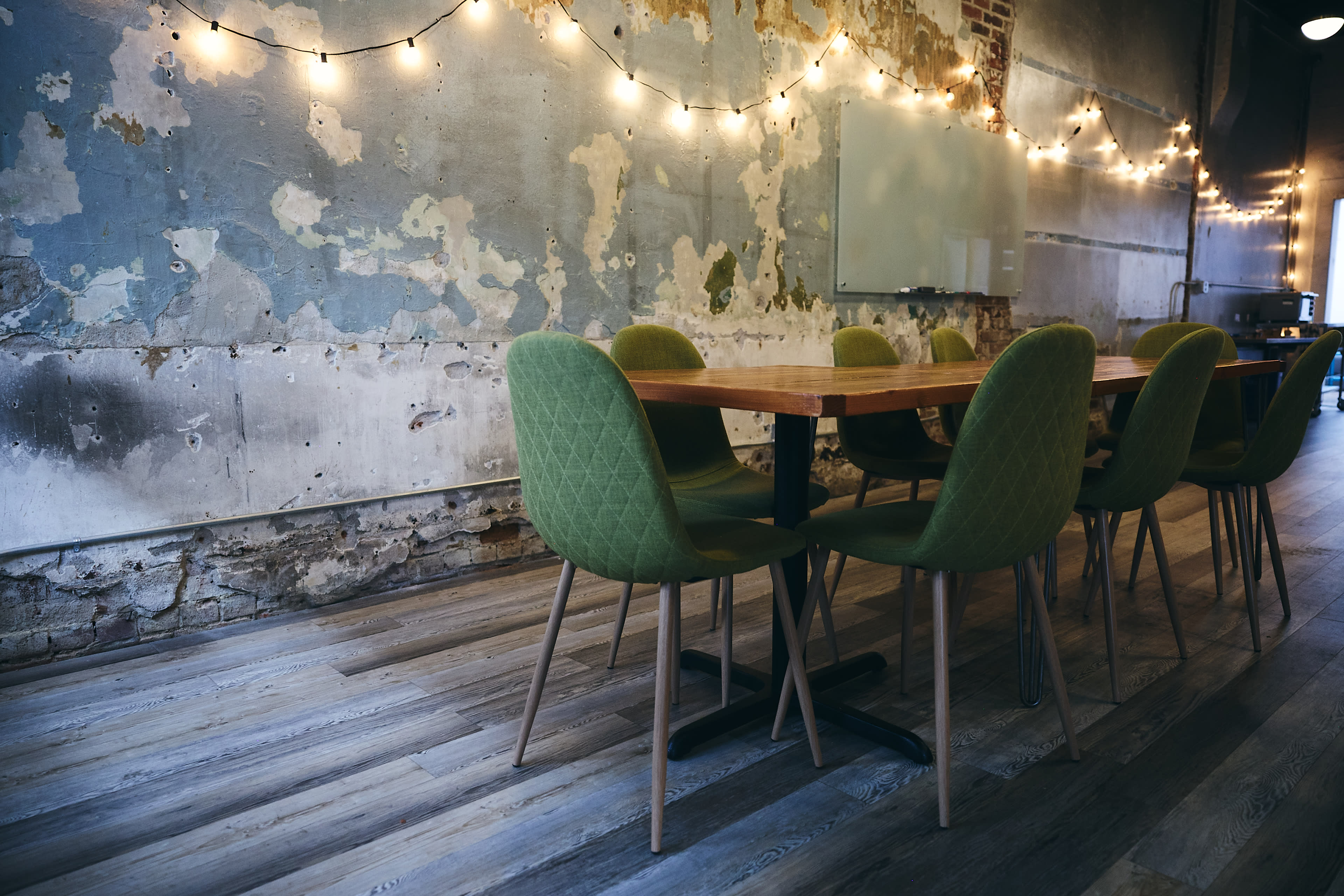 A long wooden table with green upholstered chairs is positioned in front of a distressed wall adorned with string lights.