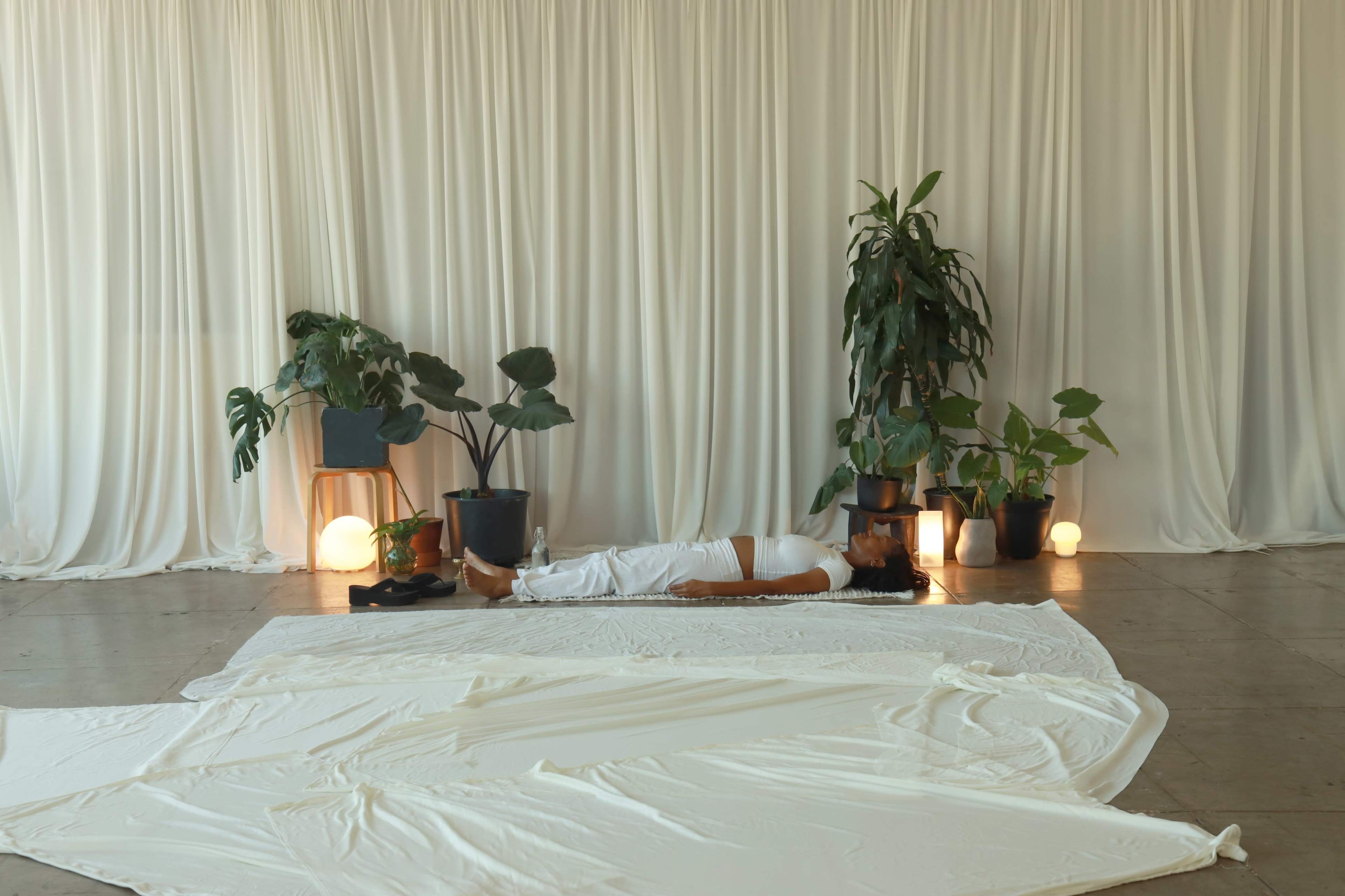 A person lies on a white mat surrounded by potted plants and soft lighting in a minimalist indoor space.