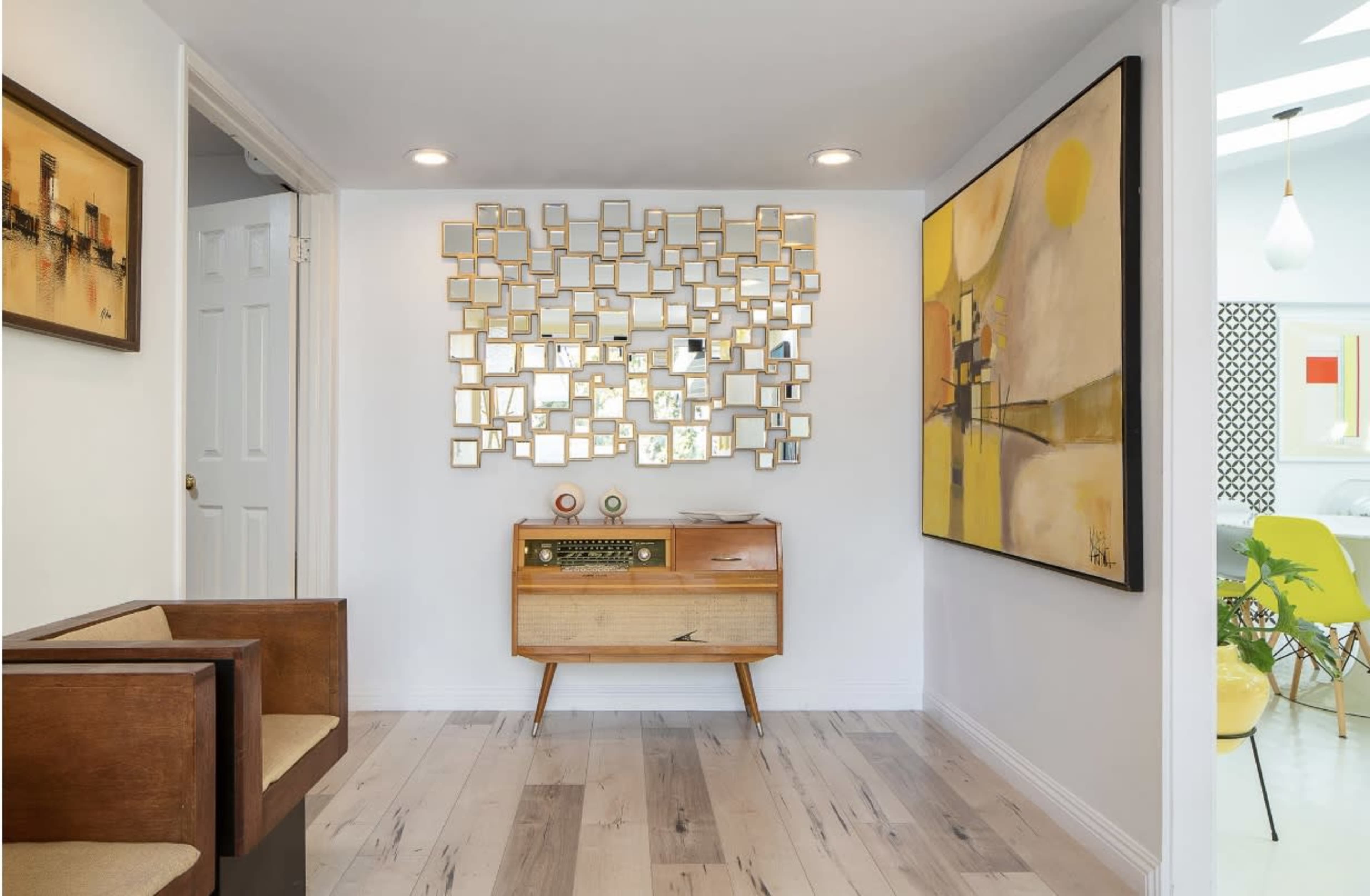 The image shows a modern hallway featuring a wall of small mirrors, a wooden console table, and a large abstract painting.