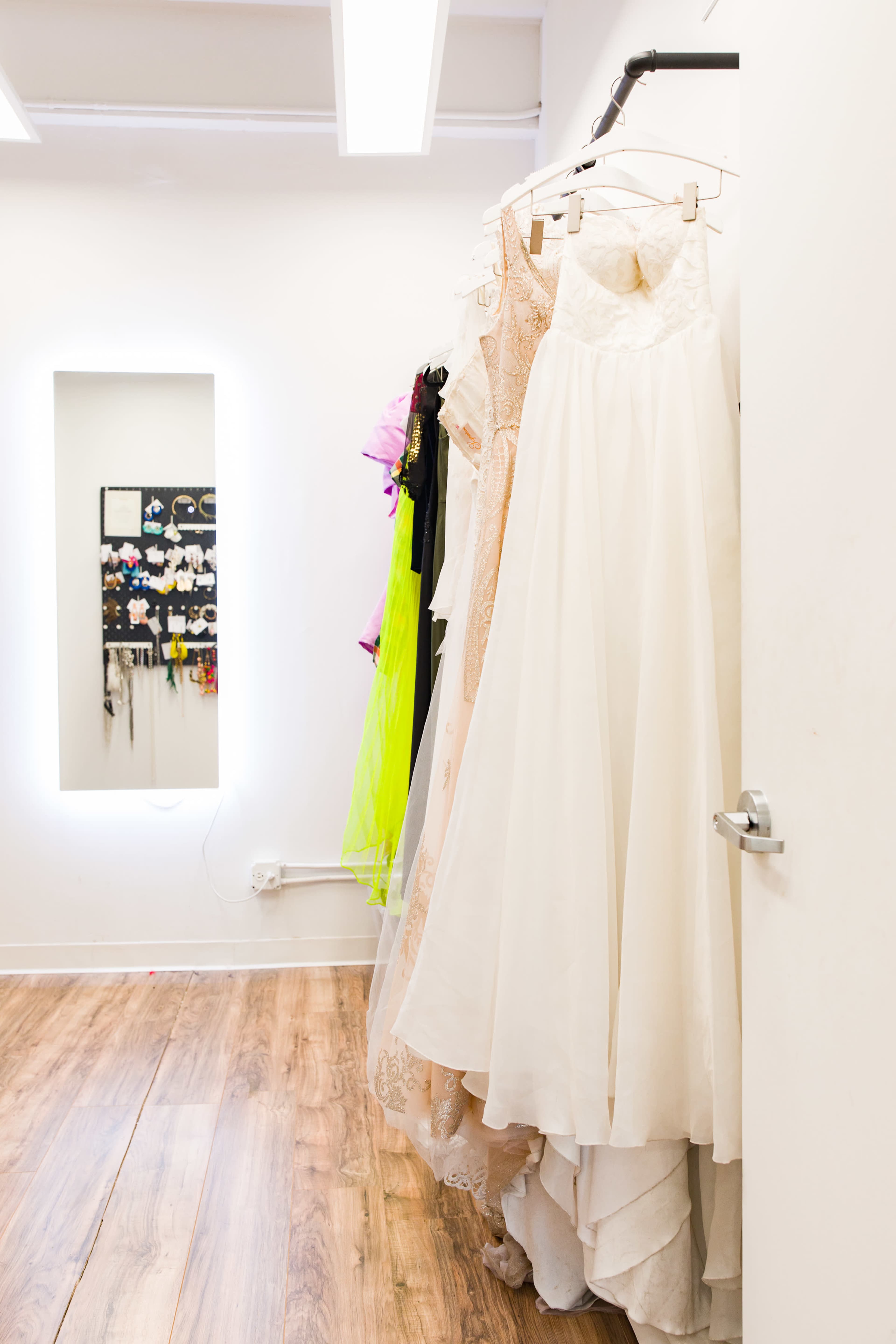 A row of dresses hangs on a clothing rack next to a mirror in a brightly lit room with wooden flooring.