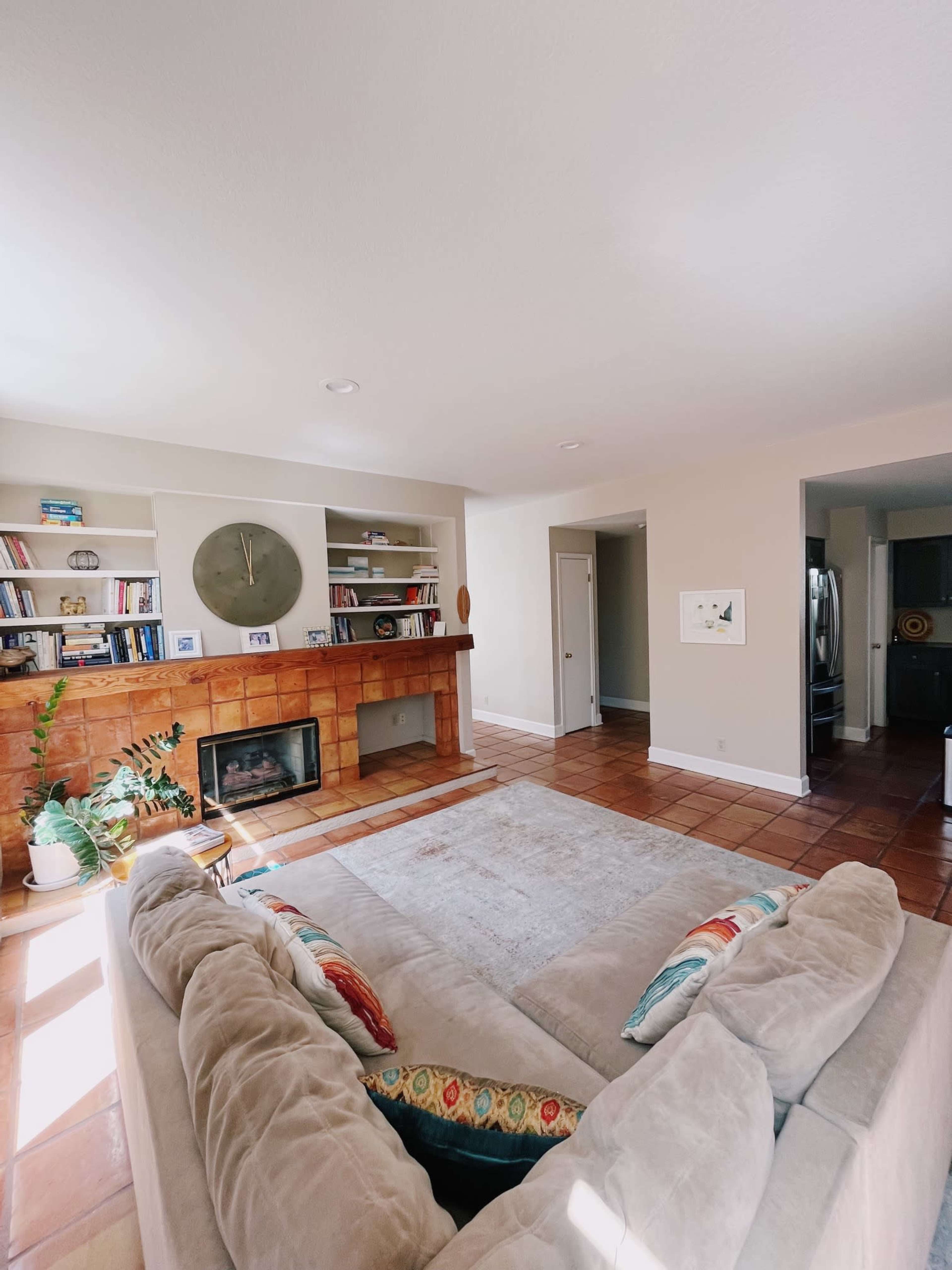The image shows a spacious living room with a beige sectional sofa, a fireplace built into a tiled wall, bookshelves, and an open doorway leading to a kitchen area.