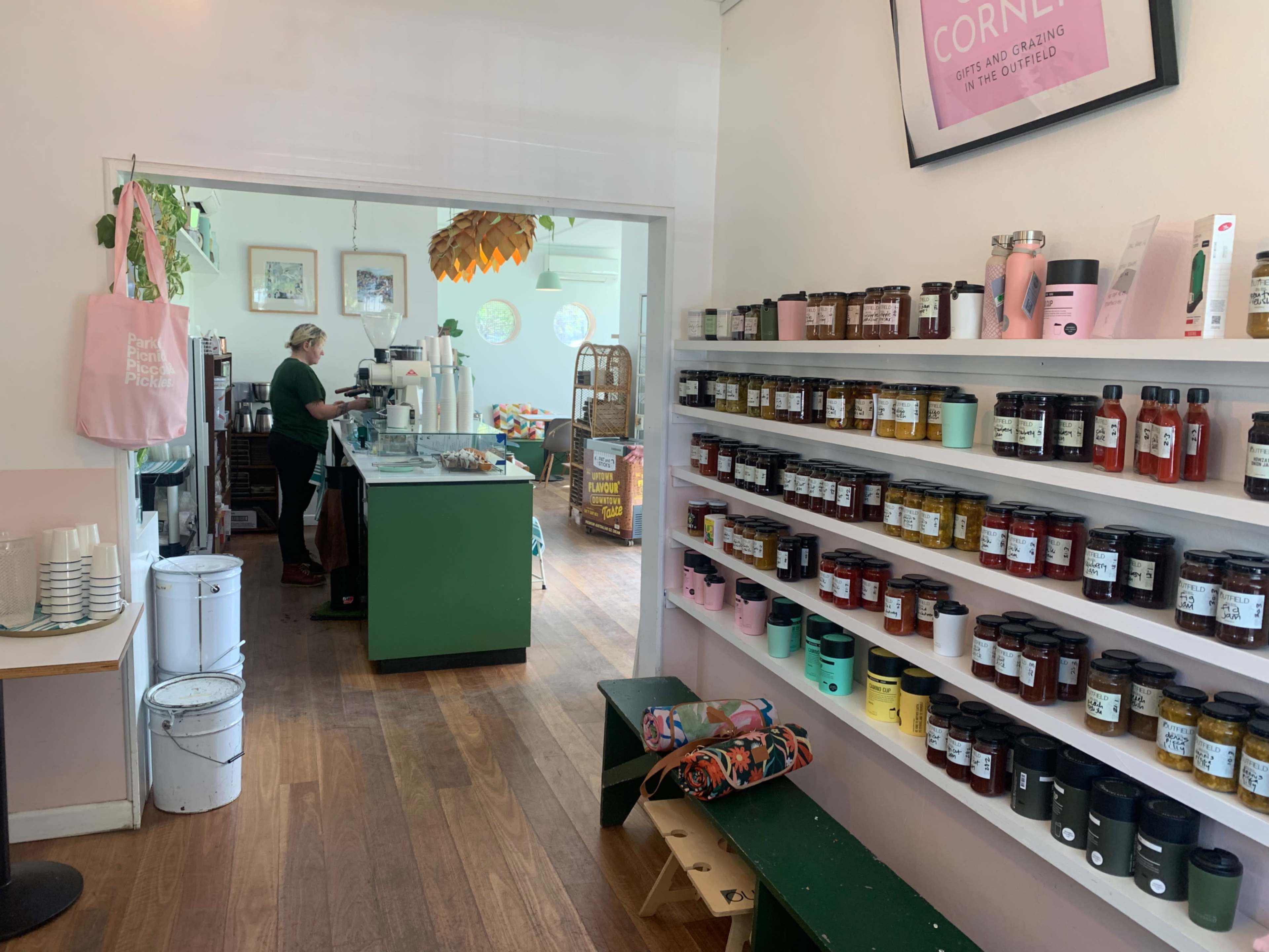 A cozy café features a counter with a barista, while shelves display jars of various jams and spreads along one wall.