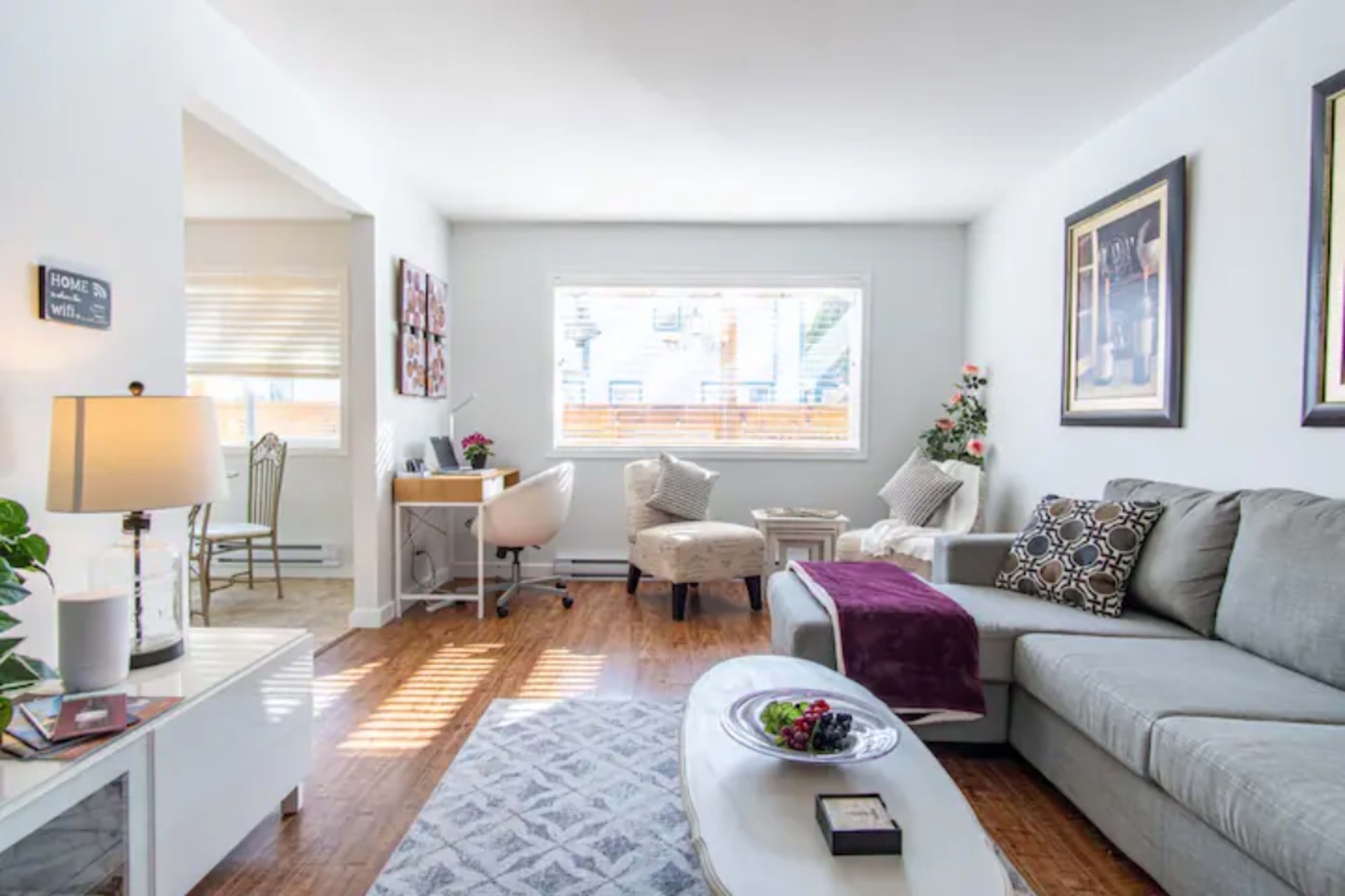 A bright living room features a gray sofa, a round white coffee table, and two armchairs, with a small desk in the corner and framed artwork on the walls.