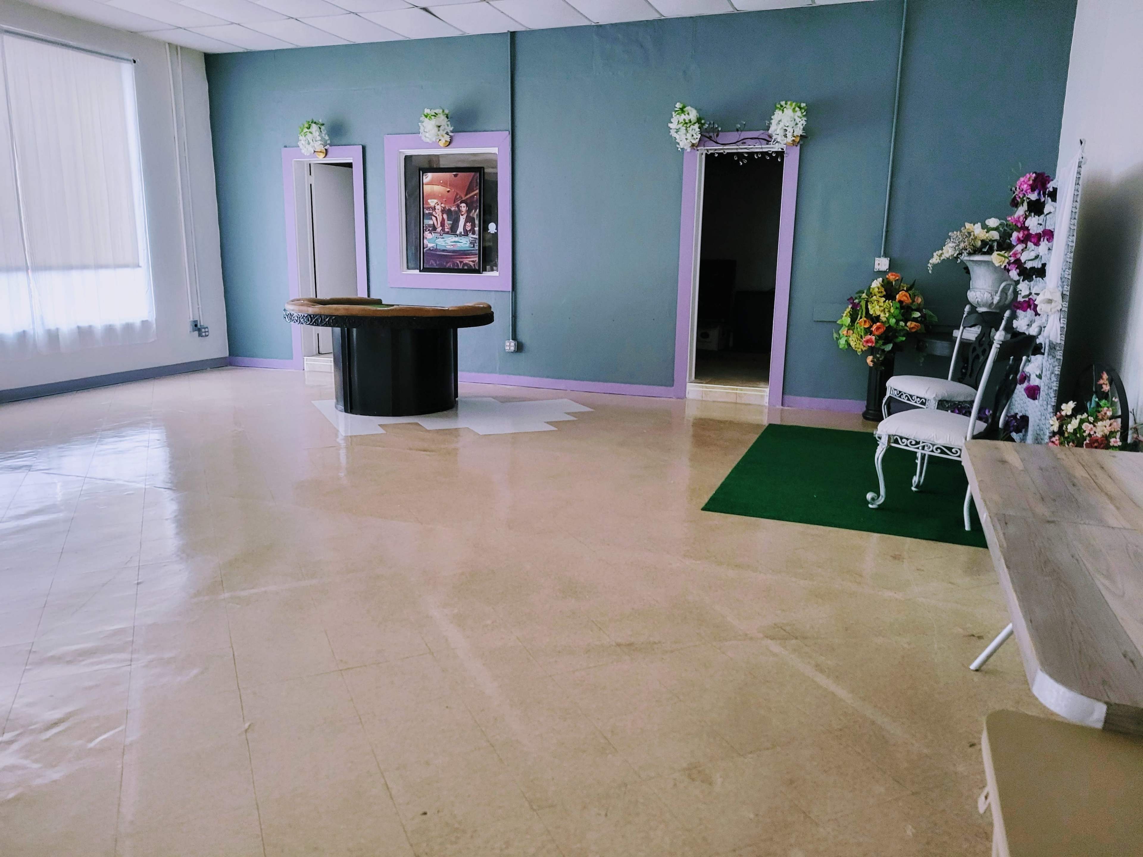 A spacious, empty room with a dark round table in the center, flanked by colorful floral arrangements and a green carpet leading to a doorway.