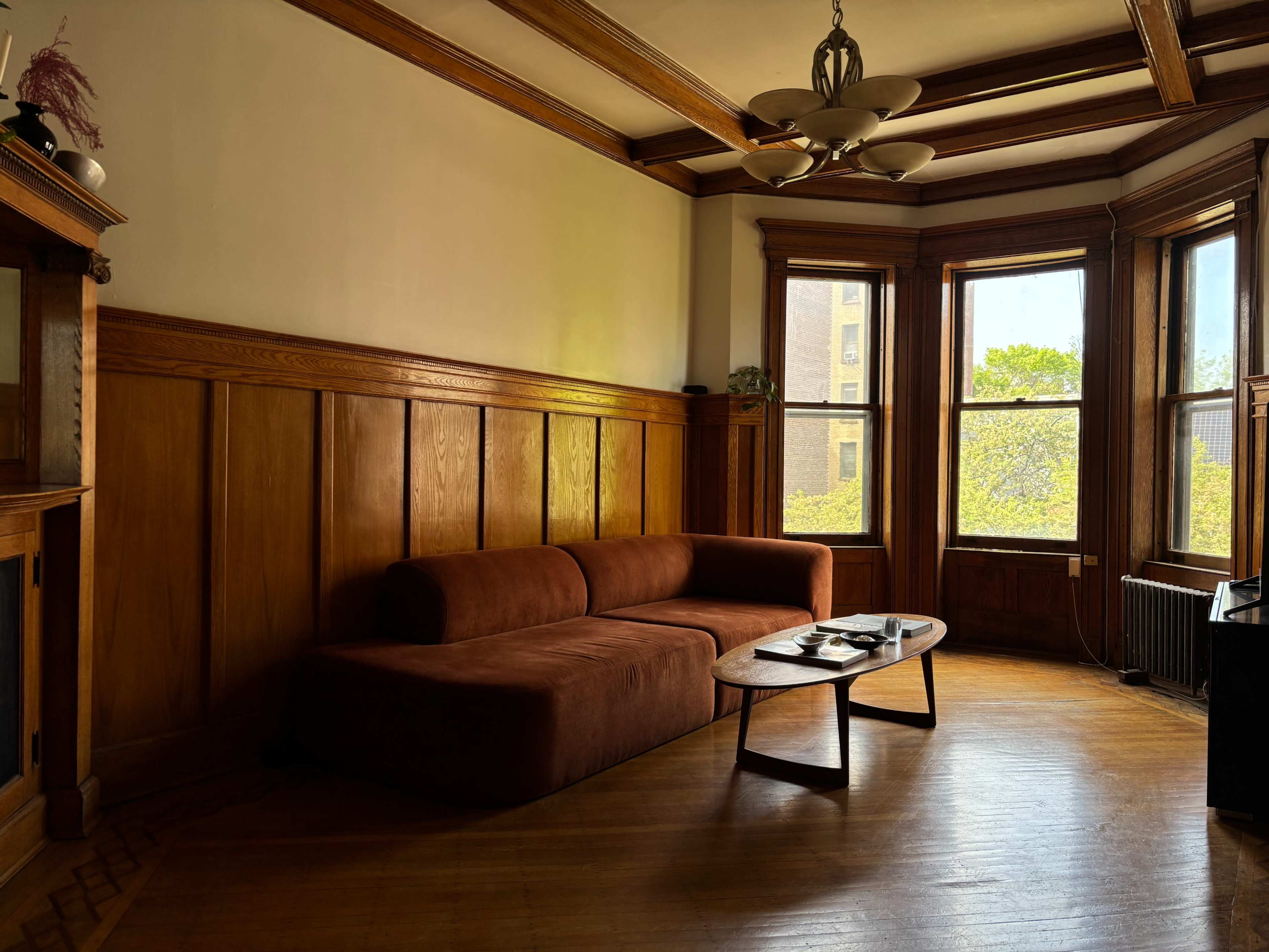 The image shows a room with wooden paneling, featuring a brown sofa and a coffee table near large windows that let in natural light.
