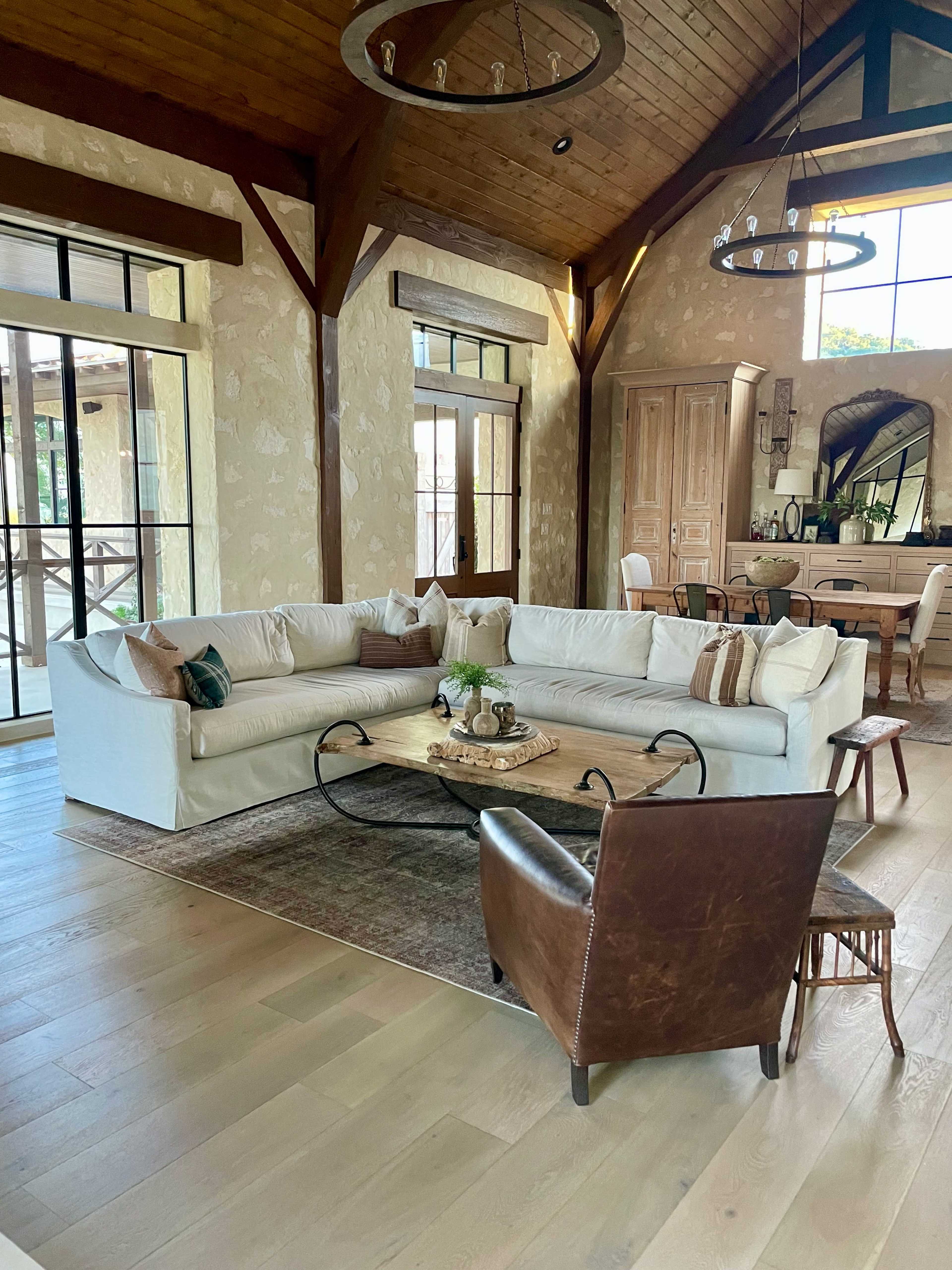 The image shows a spacious living room featuring a large white sectional sofa, a circular wooden coffee table, and a brown leather armchair, all set against a backdrop of wooden beams and large windows.