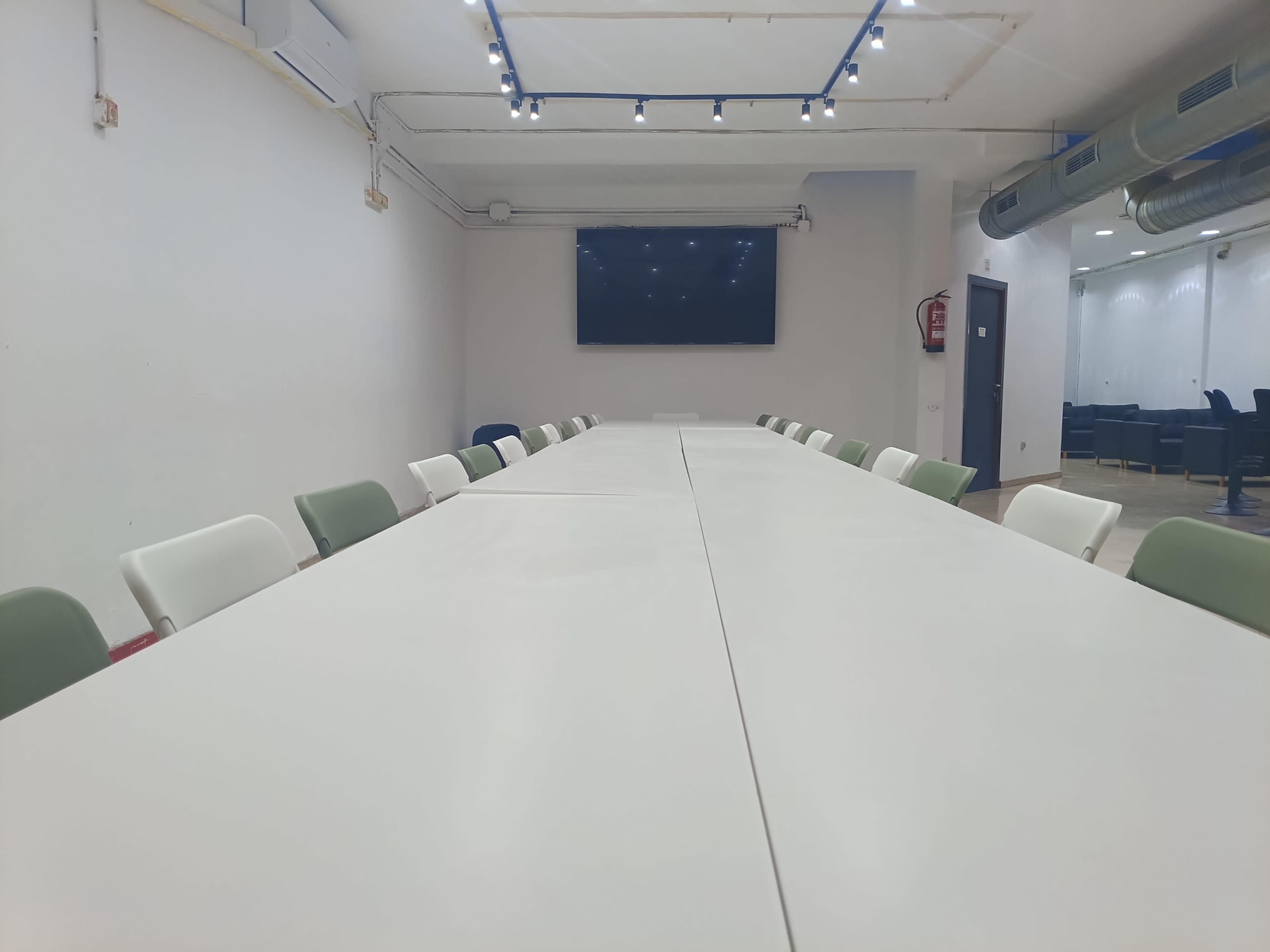 The image shows a conference room with a long white table, chairs on either side, and a wall-mounted screen at the end.