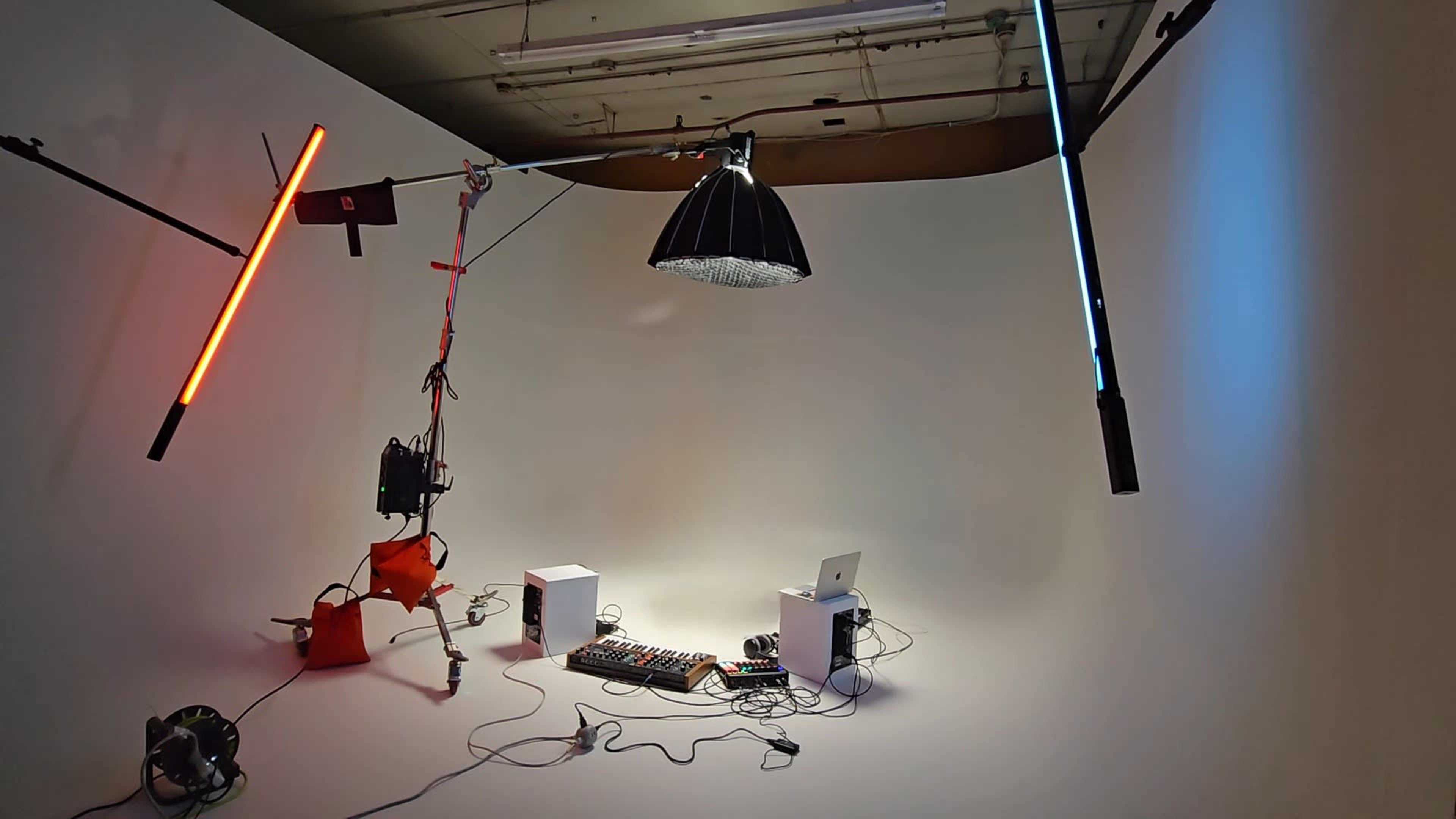 The image shows a minimalistic studio setup with colorful lighting, a music production console, and a laptop arranged against a white background.