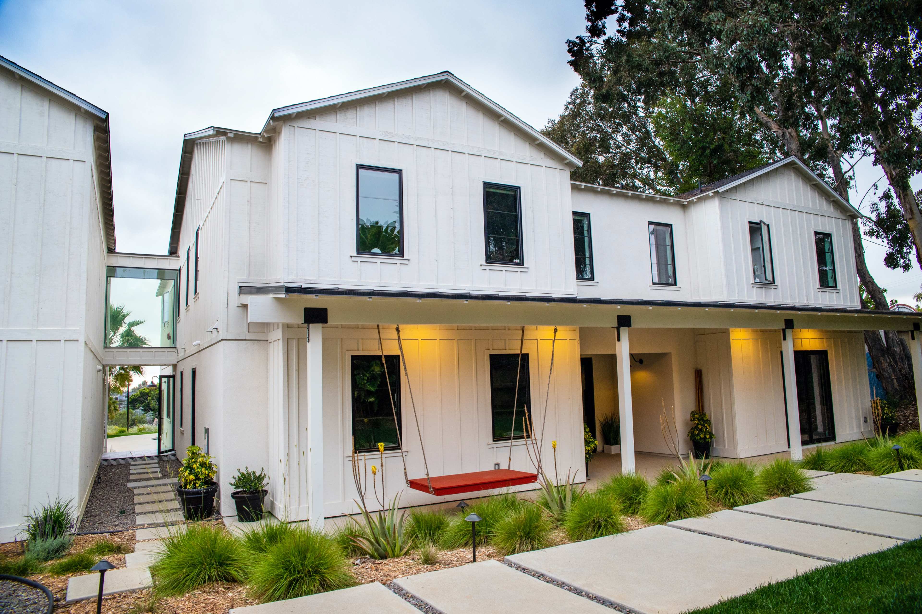 A modern, two-story white house with black windows and a porch featuring a red swing, surrounded by landscaped greenery and a pathway.
