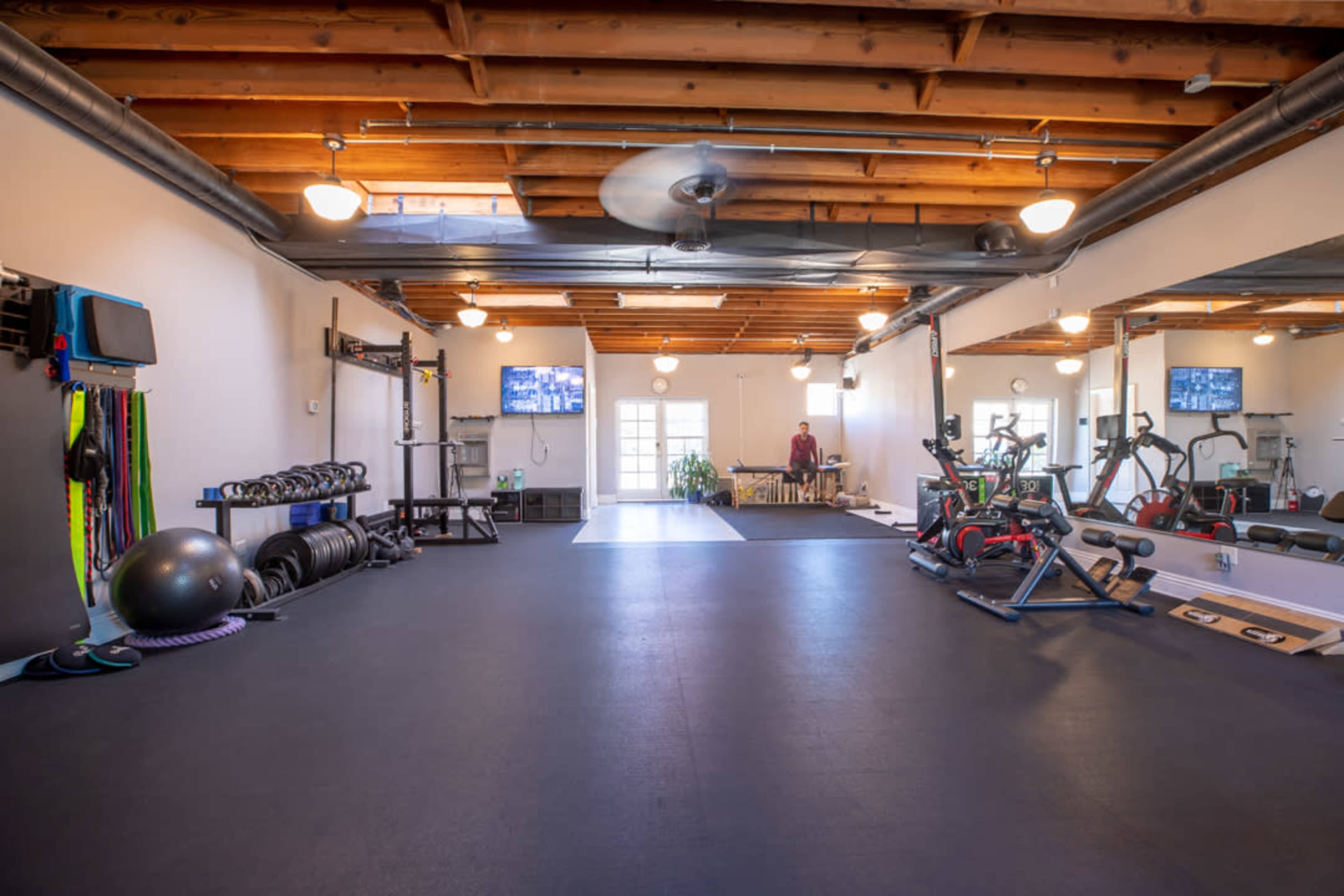 A spacious gym with exercise equipment, including weights, stationary bikes, and a wall-mounted television, beneath wooden beams and bright ceiling lights.