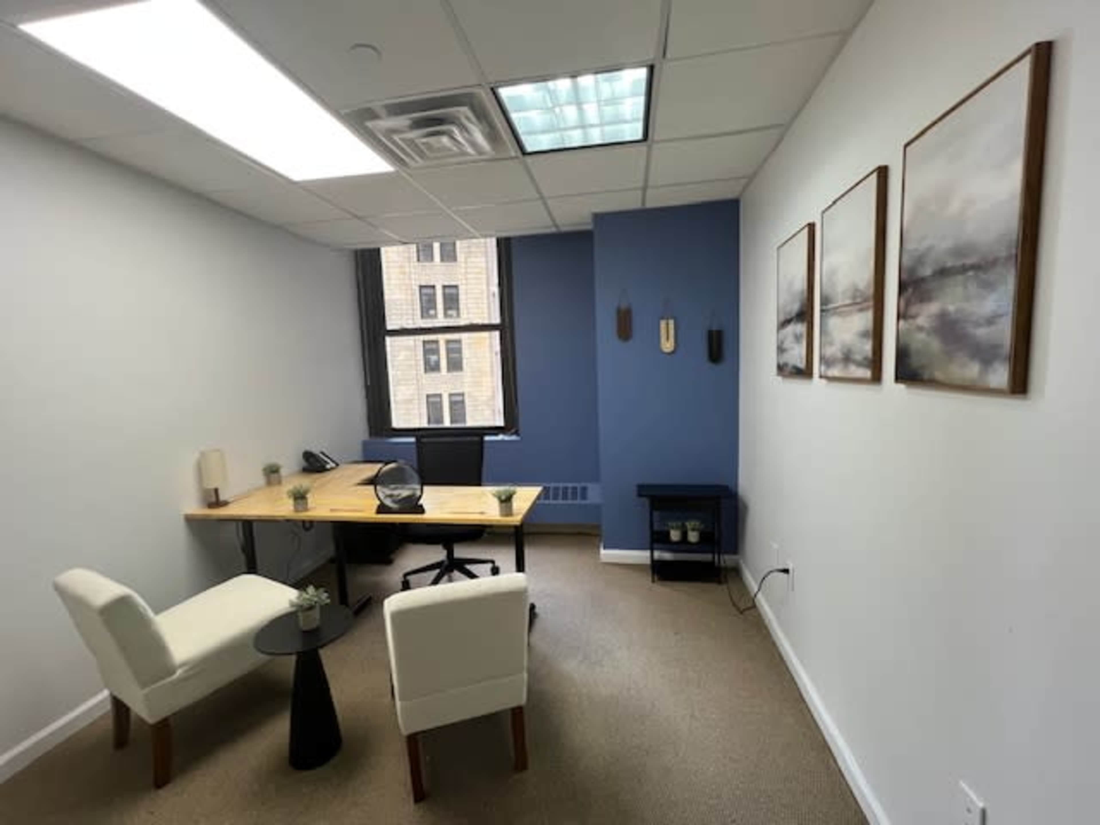 The image shows a small office with a wooden desk, two white chairs, and three framed pictures on the wall, along with a window providing natural light.