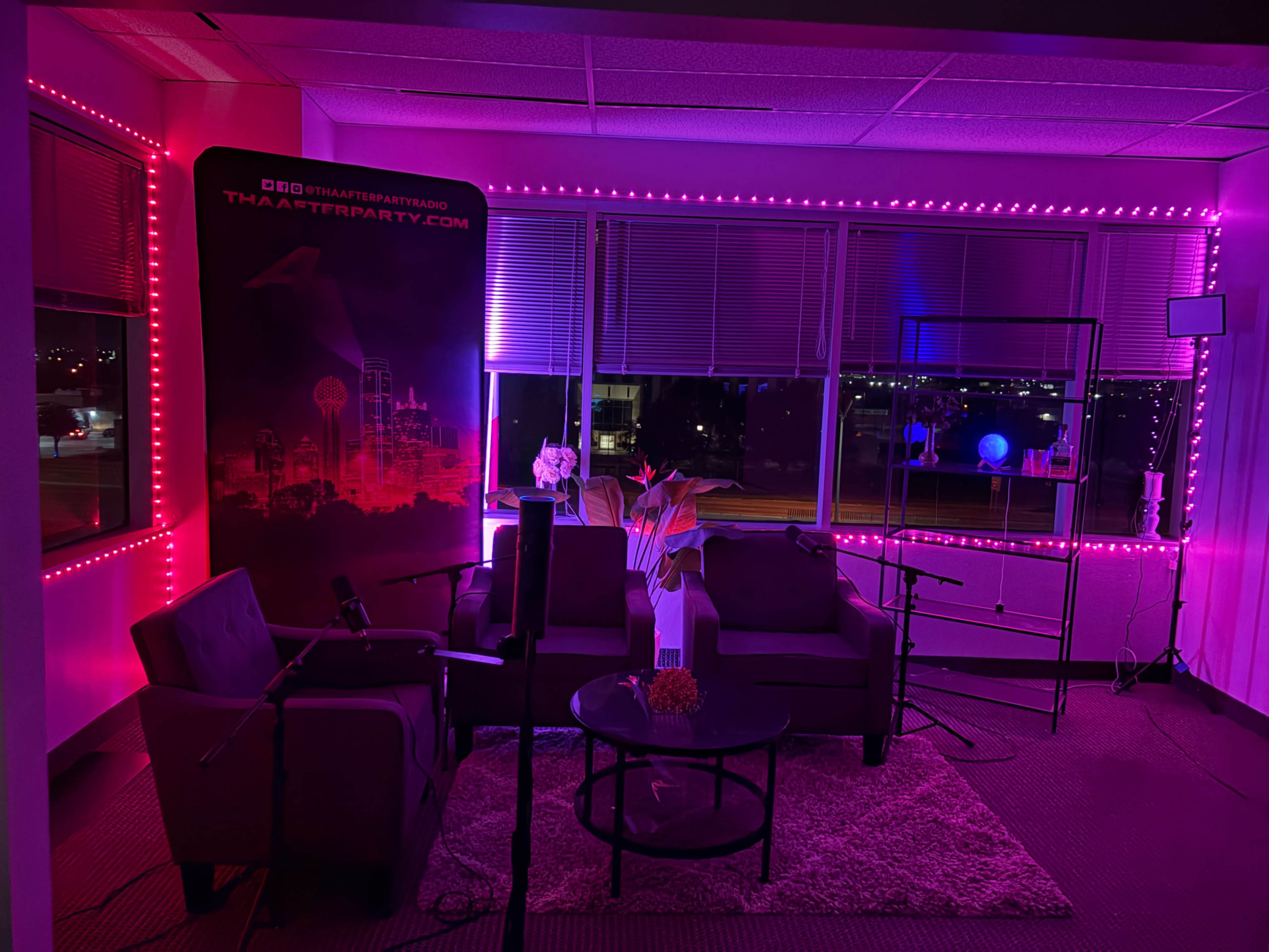 The image shows a modern, brightly lit studio space with pink LED lights, featuring a seating area with two chairs, a coffee table, and city views through large windows.