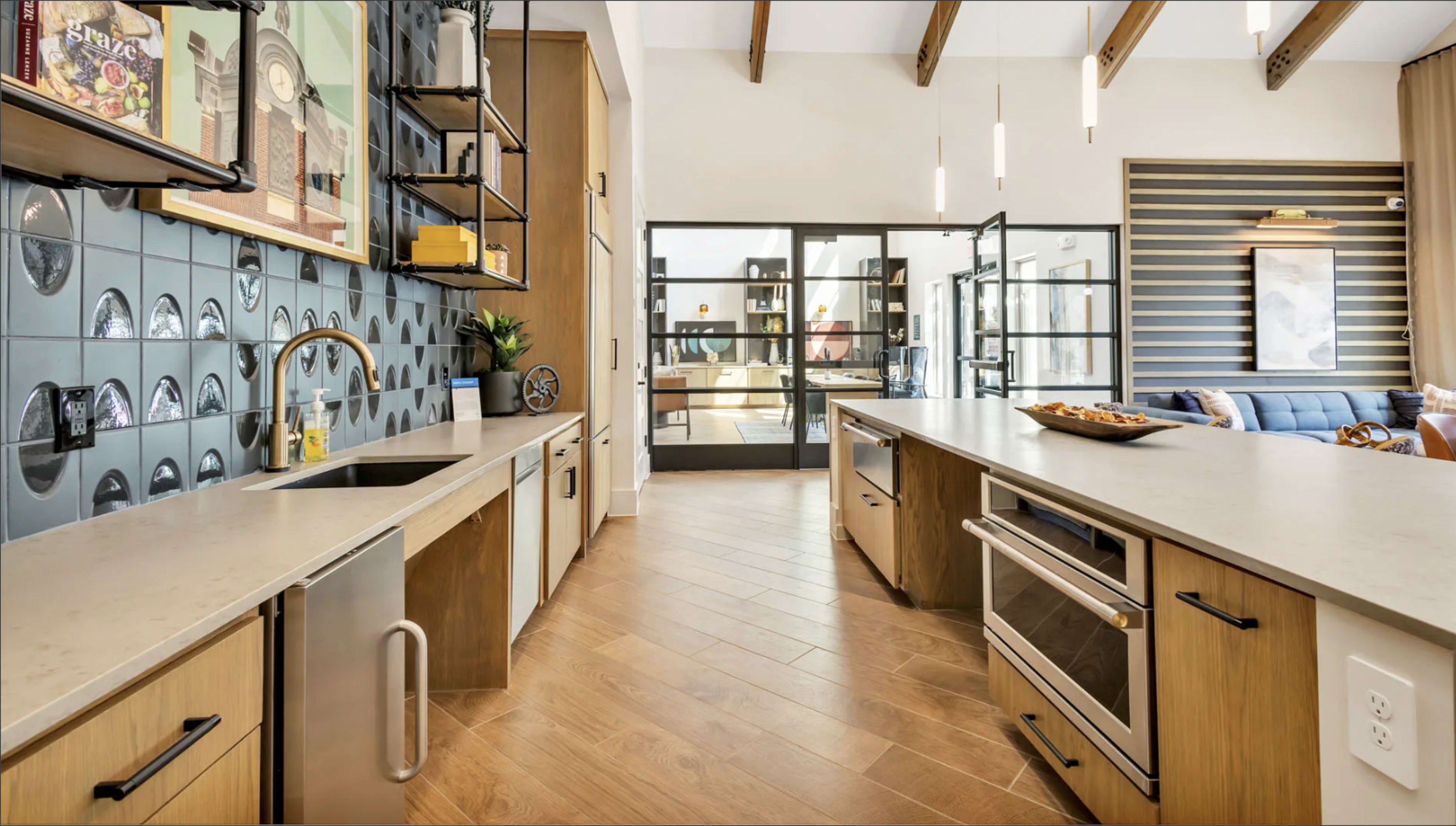 The image shows a modern kitchen featuring wooden cabinets, a large island with a countertop, and a view into a living area through large glass doors.