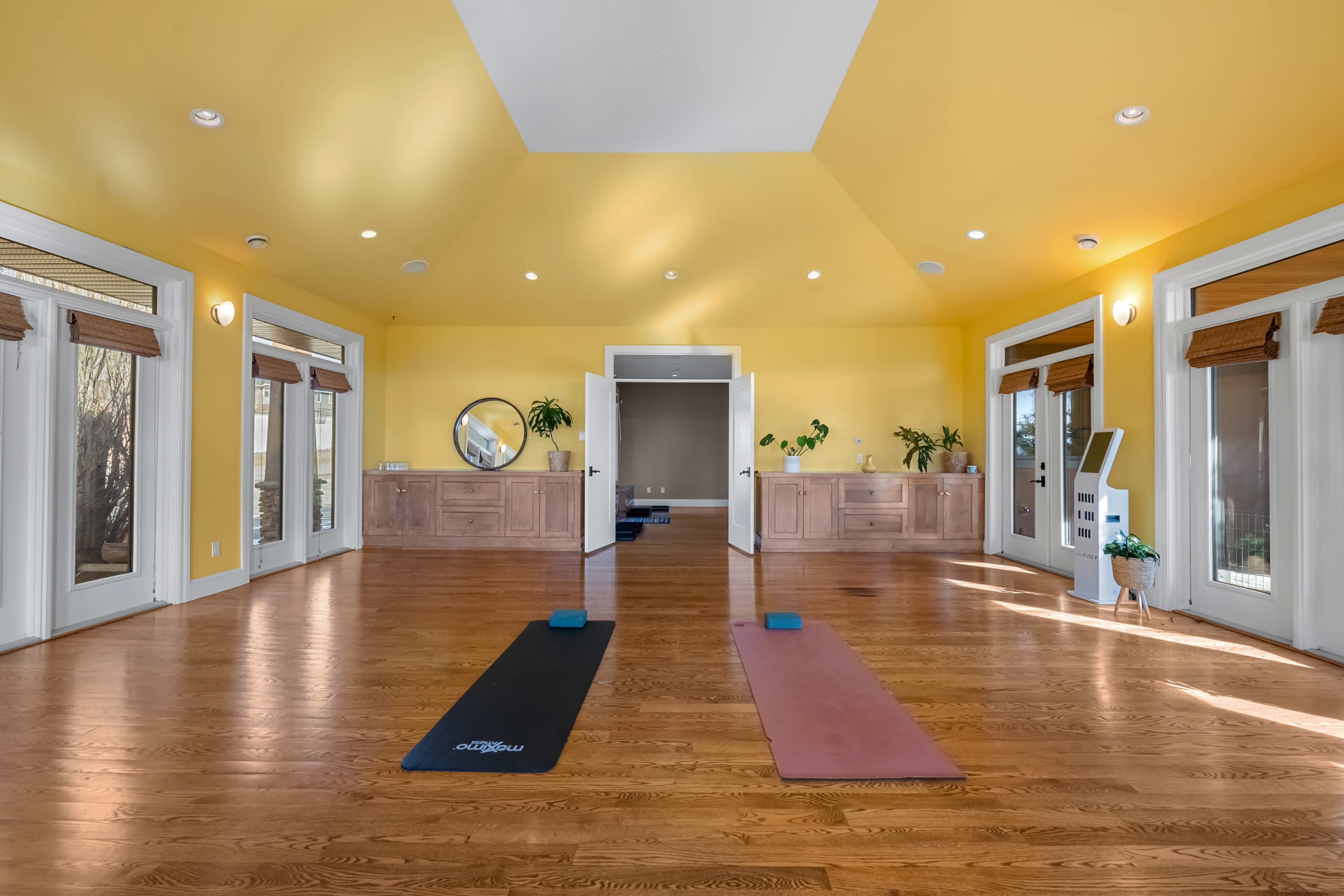 The image shows a bright, spacious yoga studio with two yoga mats on a wooden floor and large windows around the room.