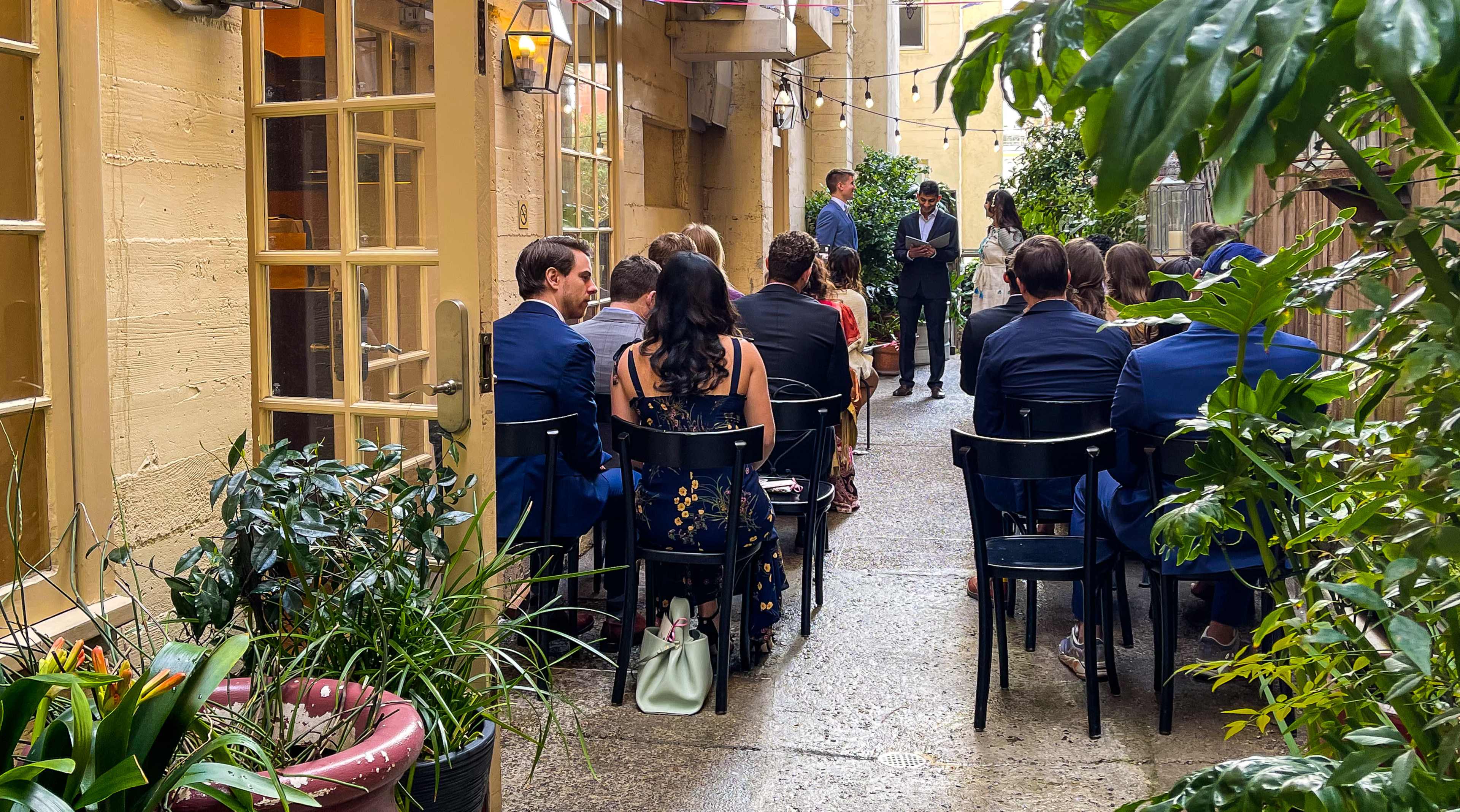 A group of people is seated in a narrow outdoor space lined with plants, while two individuals stand at the front, likely giving a speech or presentation.