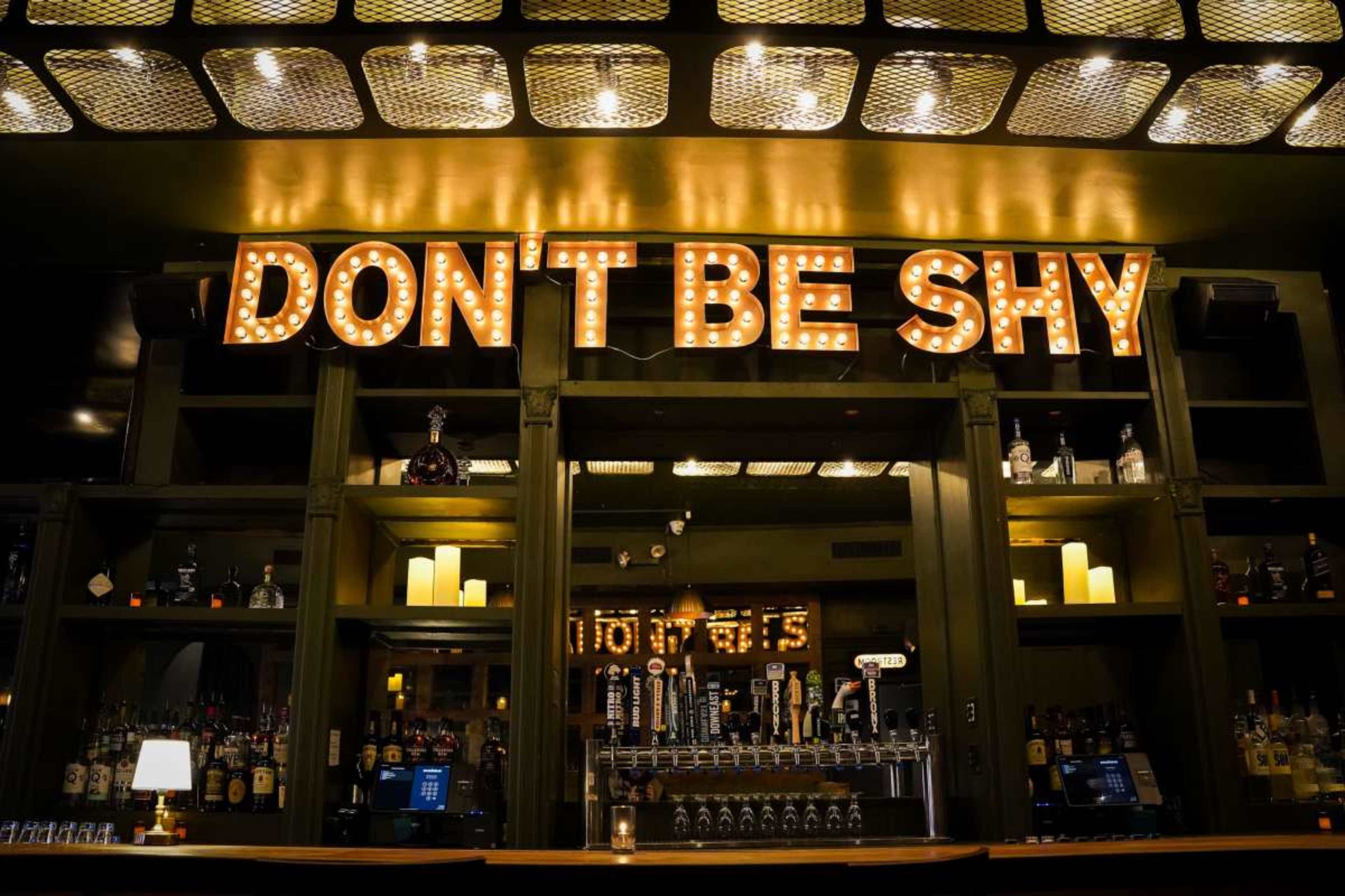 The image features a bar with vintage-style illuminated letters spelling "DON'T BE SHY" above shelves lined with various bottles and a row of taps.
