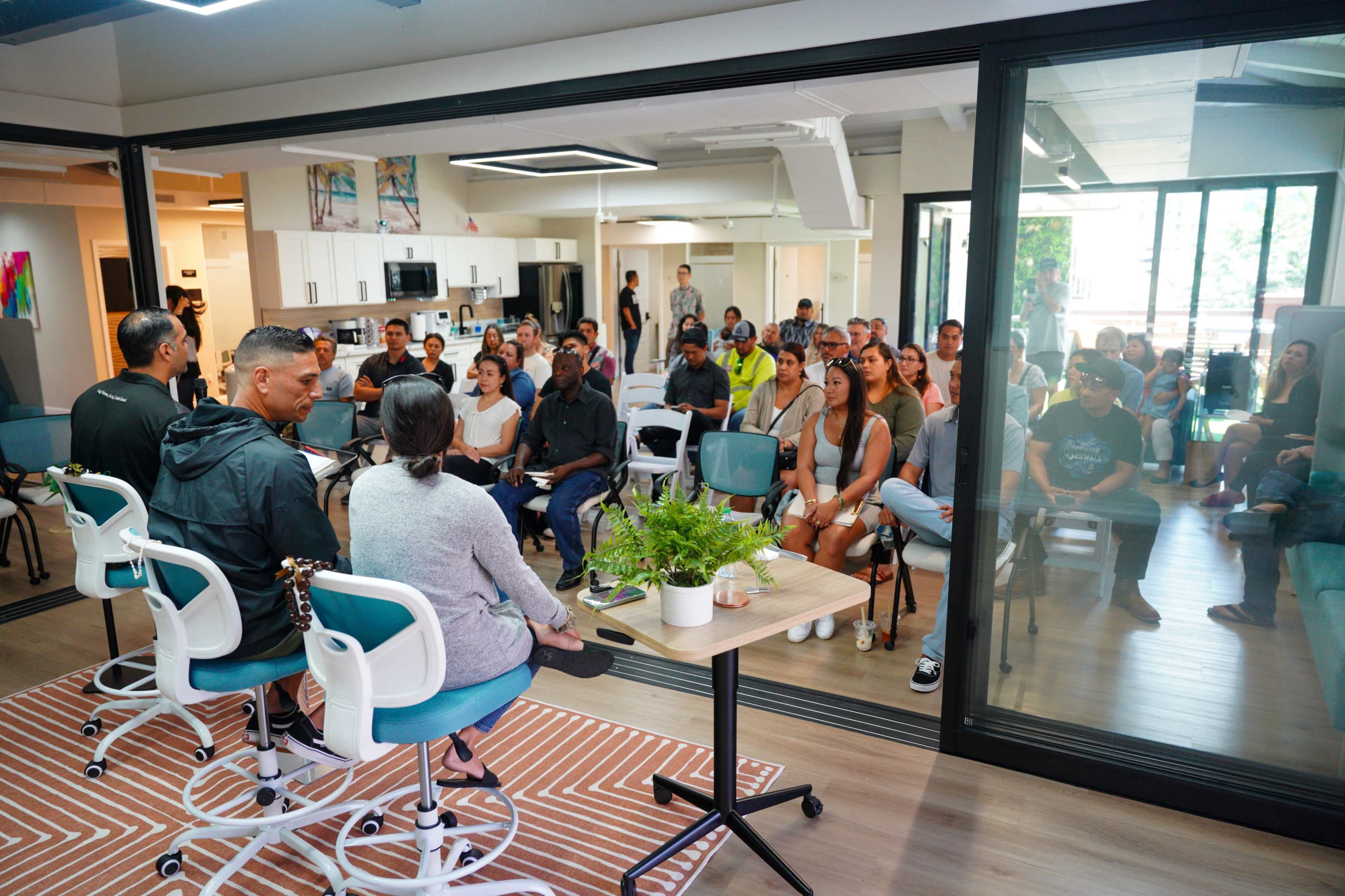 A group of people sits attentively in a modern meeting space, facing two speakers at the front.
