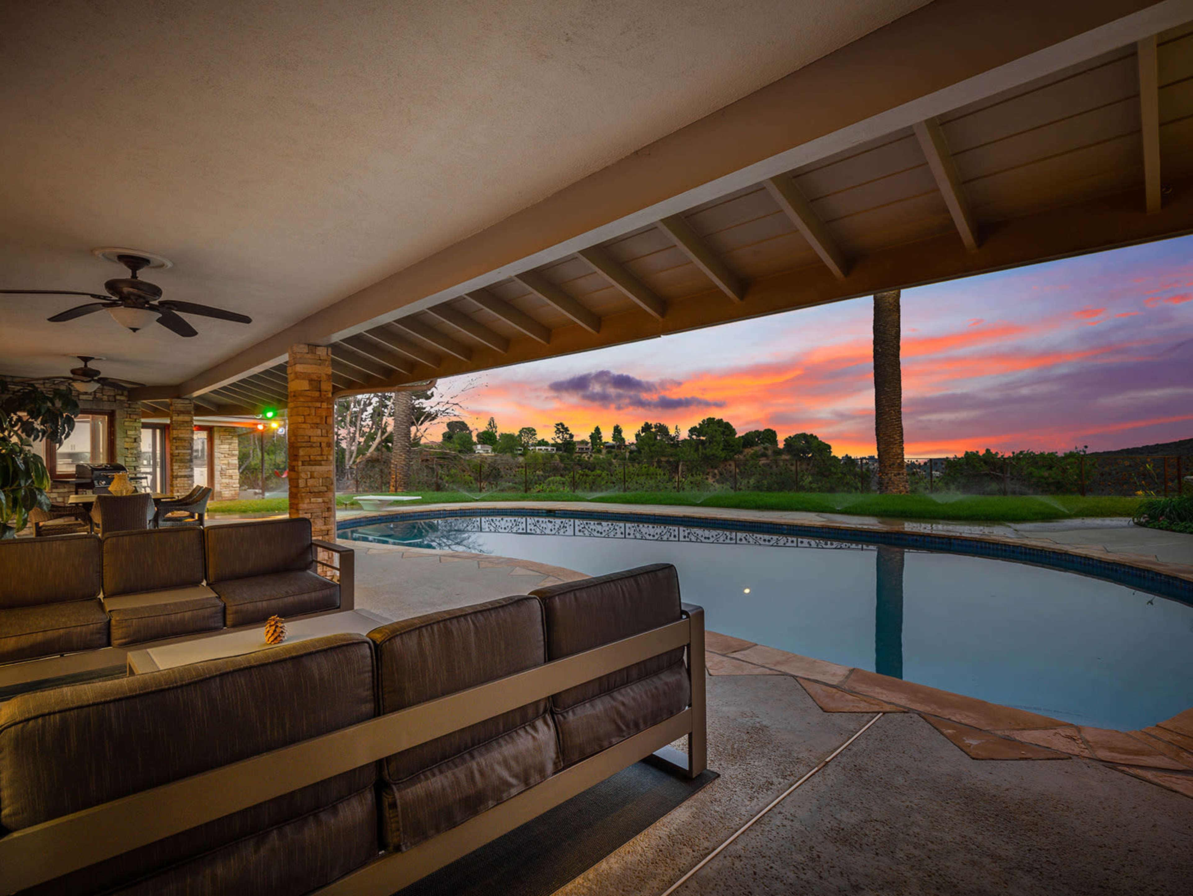 A covered patio area features a sectional couch beside a swimming pool, overlooking a colorful sunset.