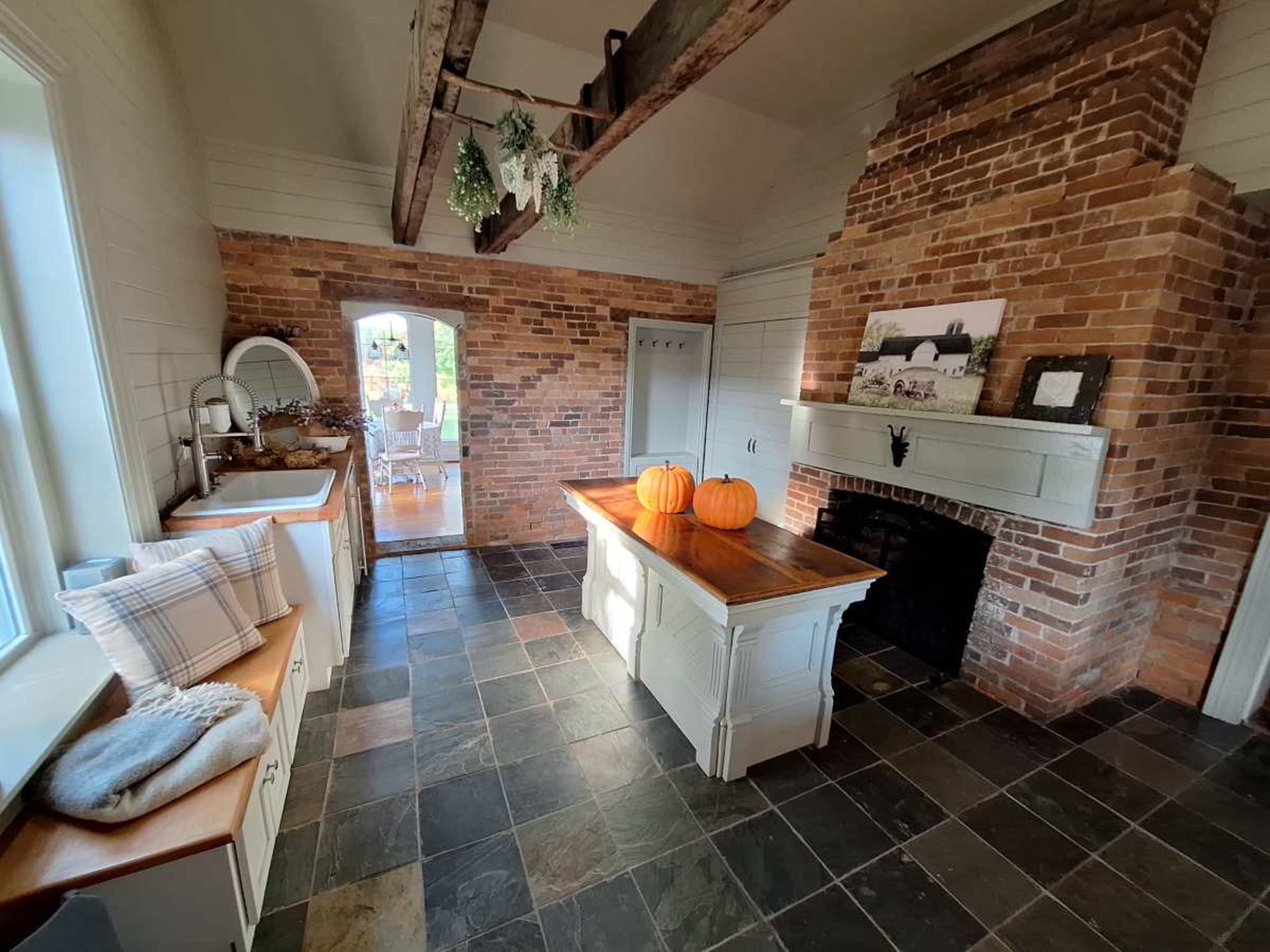The image shows a rustic kitchen with exposed brick walls, a central wooden island, and three pumpkins on the counter.