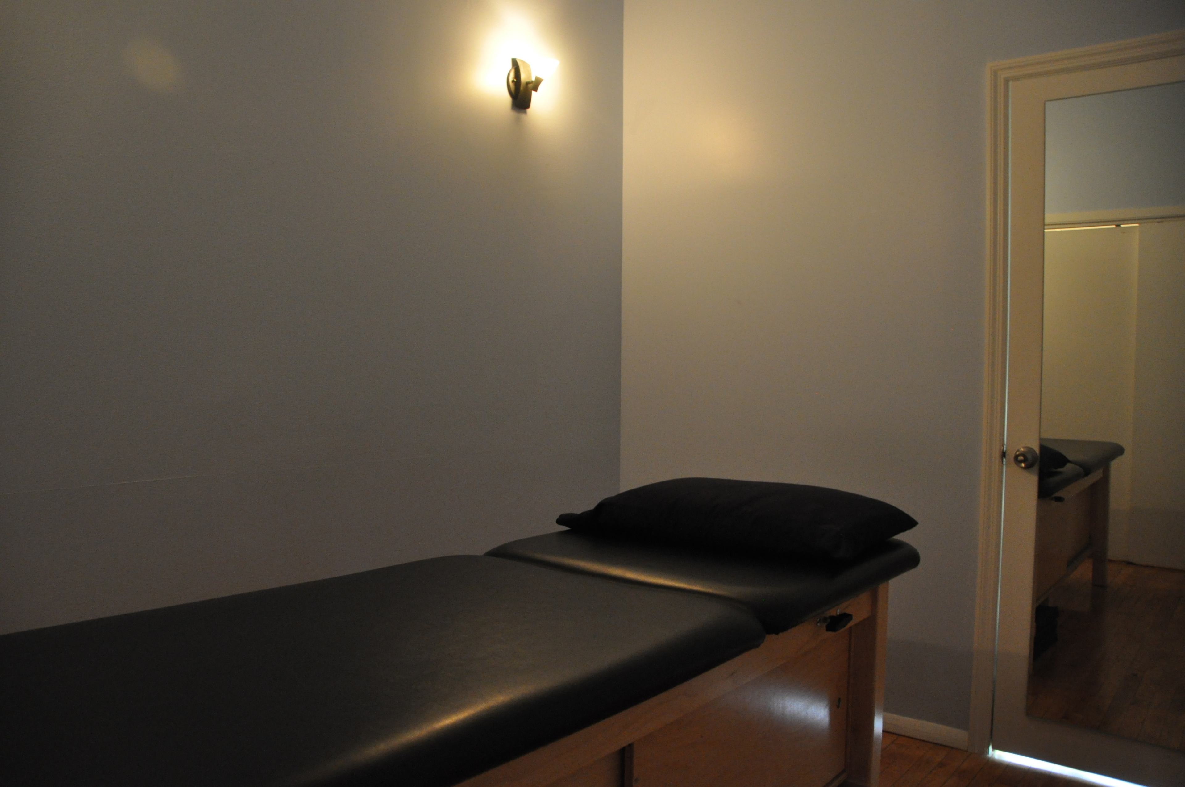 A small treatment room with a massage table, a black pillow on the table, and a mirror reflecting the room's interior.