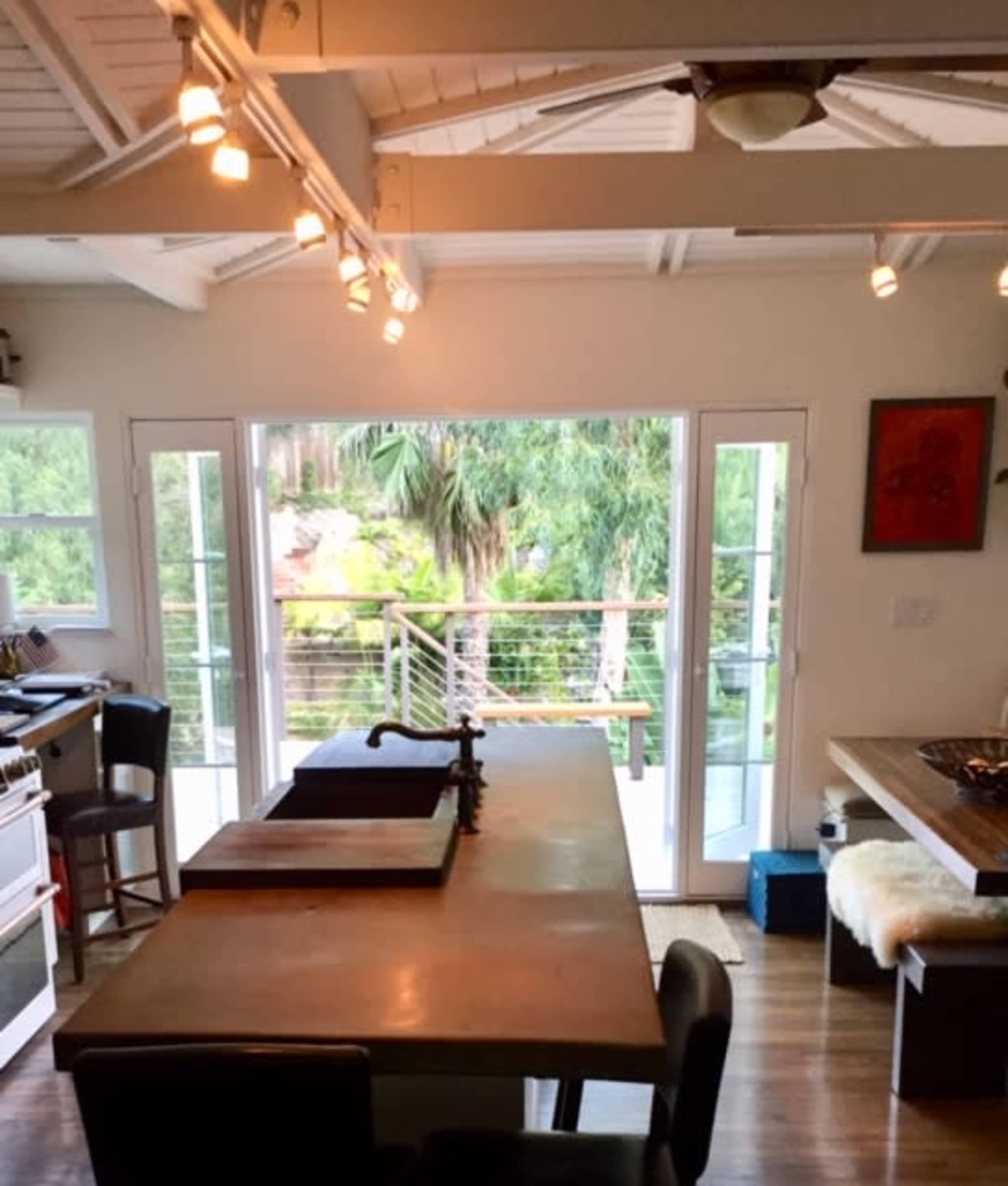 A modern kitchen features an island with a sink, large windows overlooking a patio with palm trees, and sliding glass doors leading outside.
