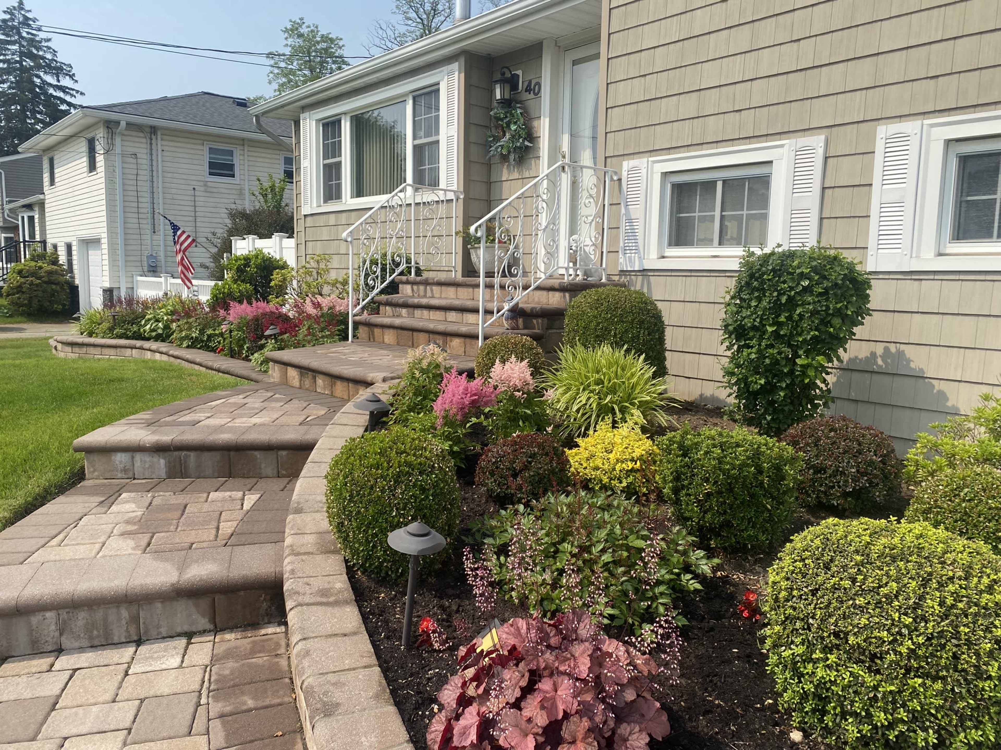A well-maintained front yard with a stone pathway leading to a house, surrounded by various colorful plants and neatly trimmed hedges.