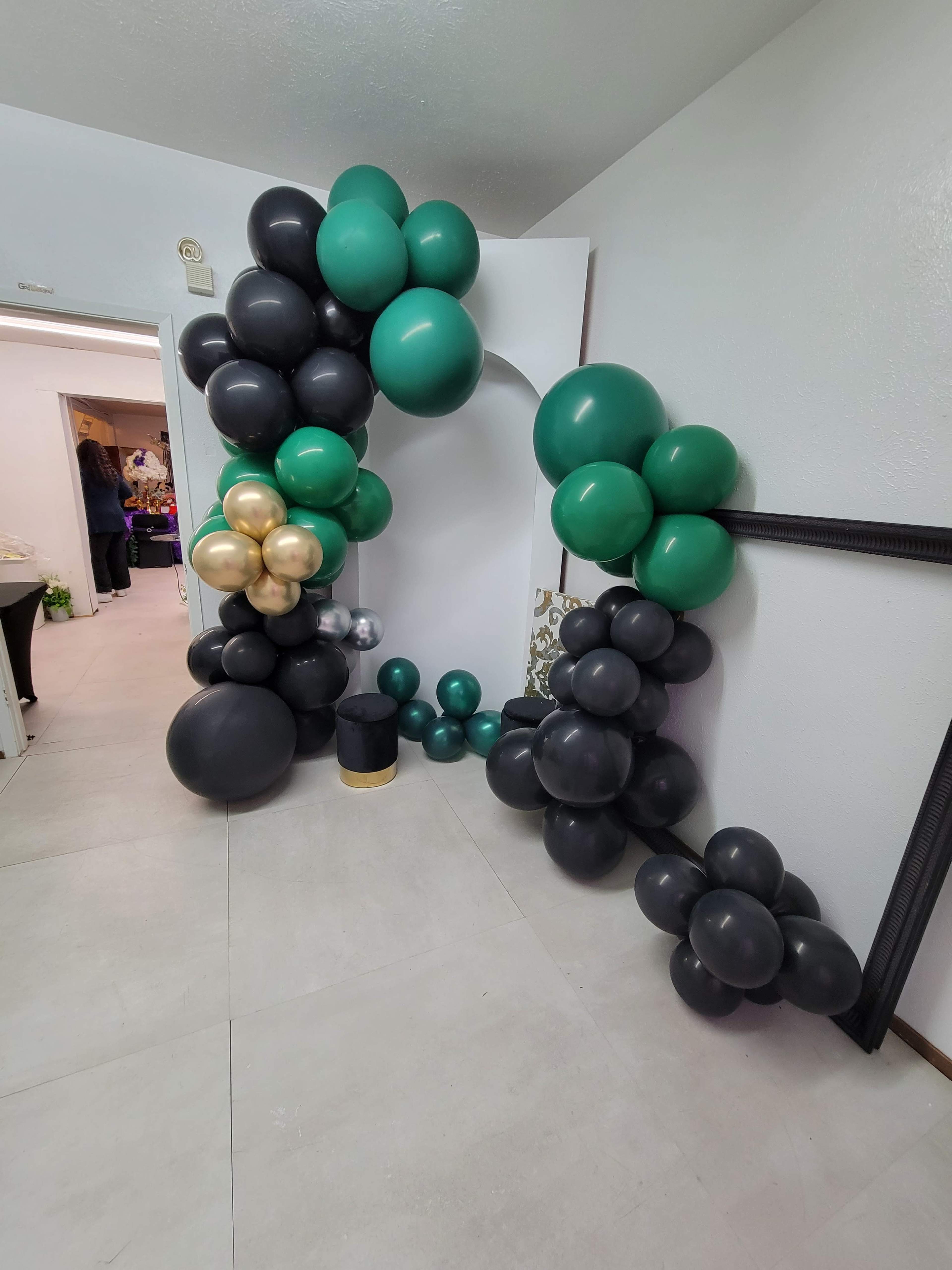 A decorative arch made of green, black, and gold balloons in a room with white walls and tile flooring.