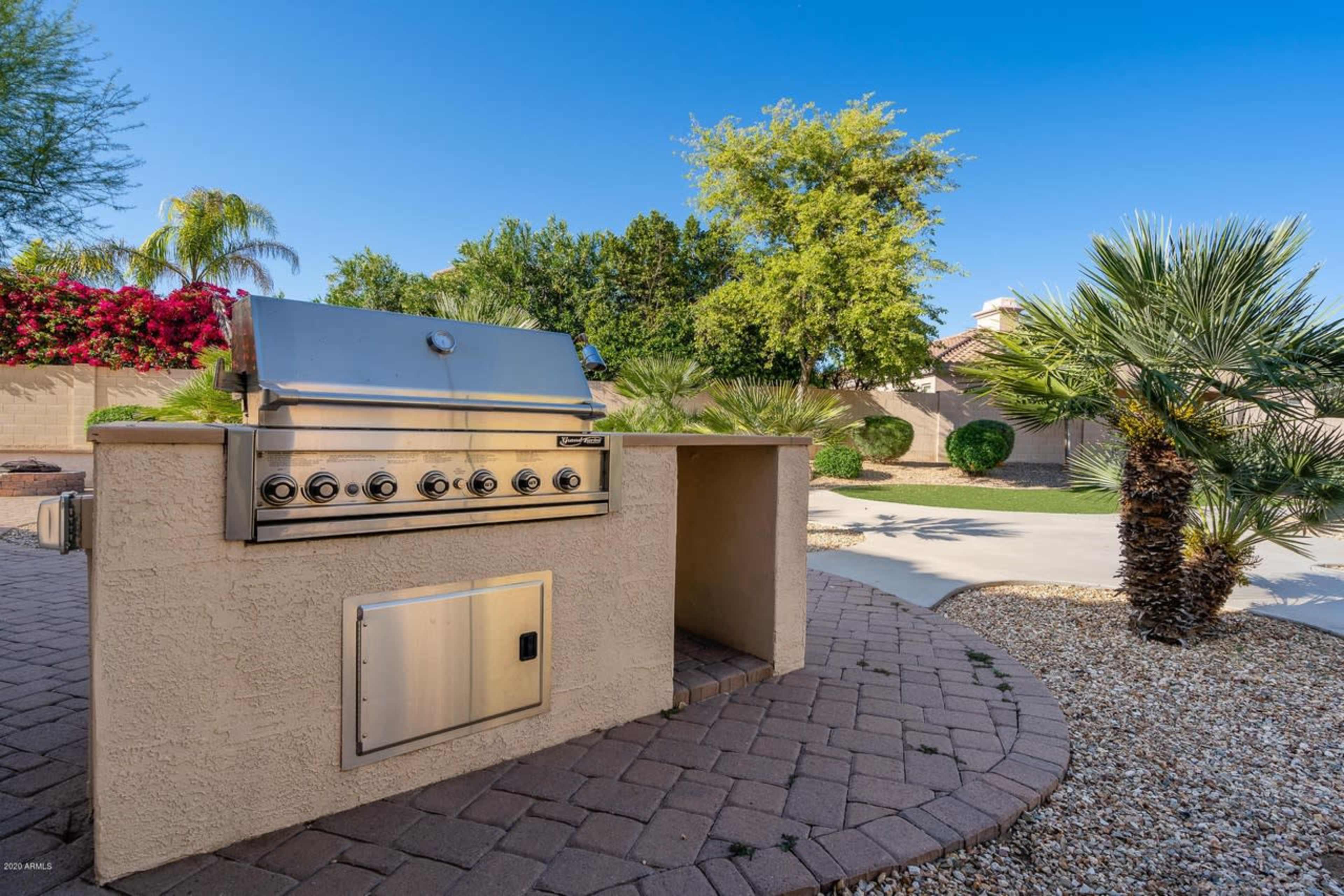 The image shows an outdoor grilling station made of stone with a stainless steel grill situated in a landscaped backyard.