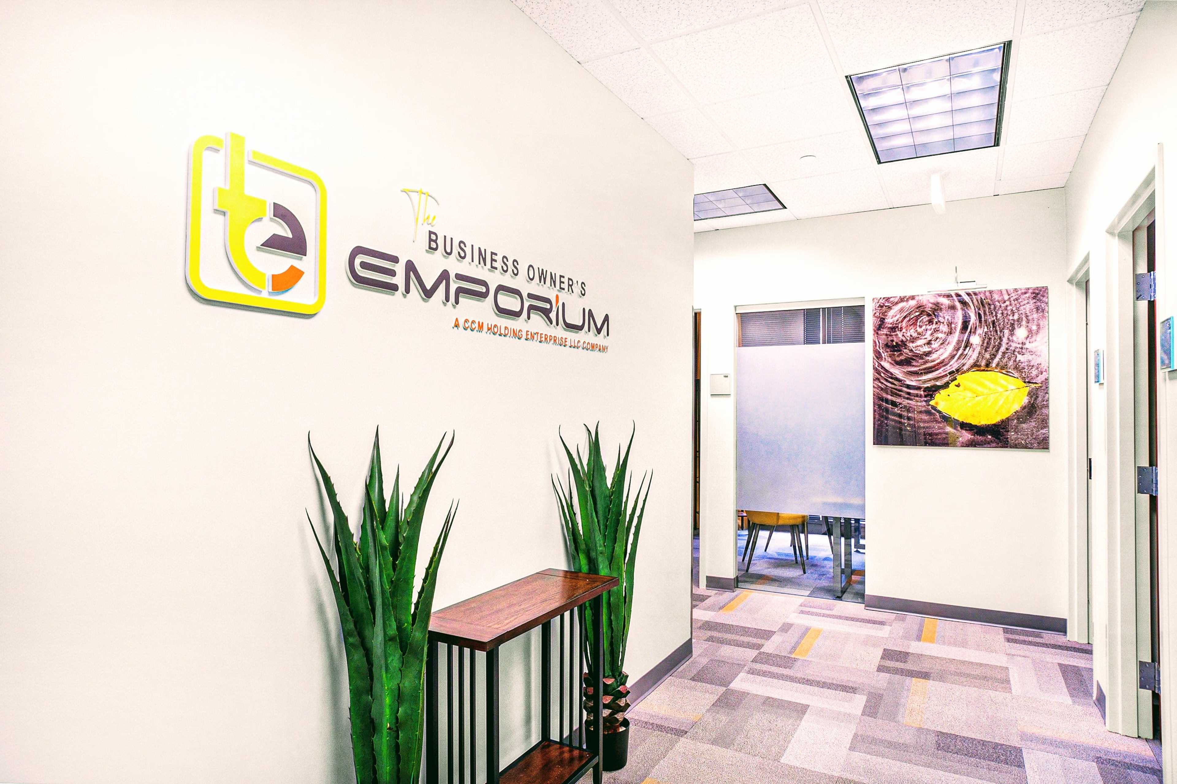 A well-lit, modern office hallway featuring a sign for "The Business Owner's Emporium" and decorative plants alongside a wooden shelf.