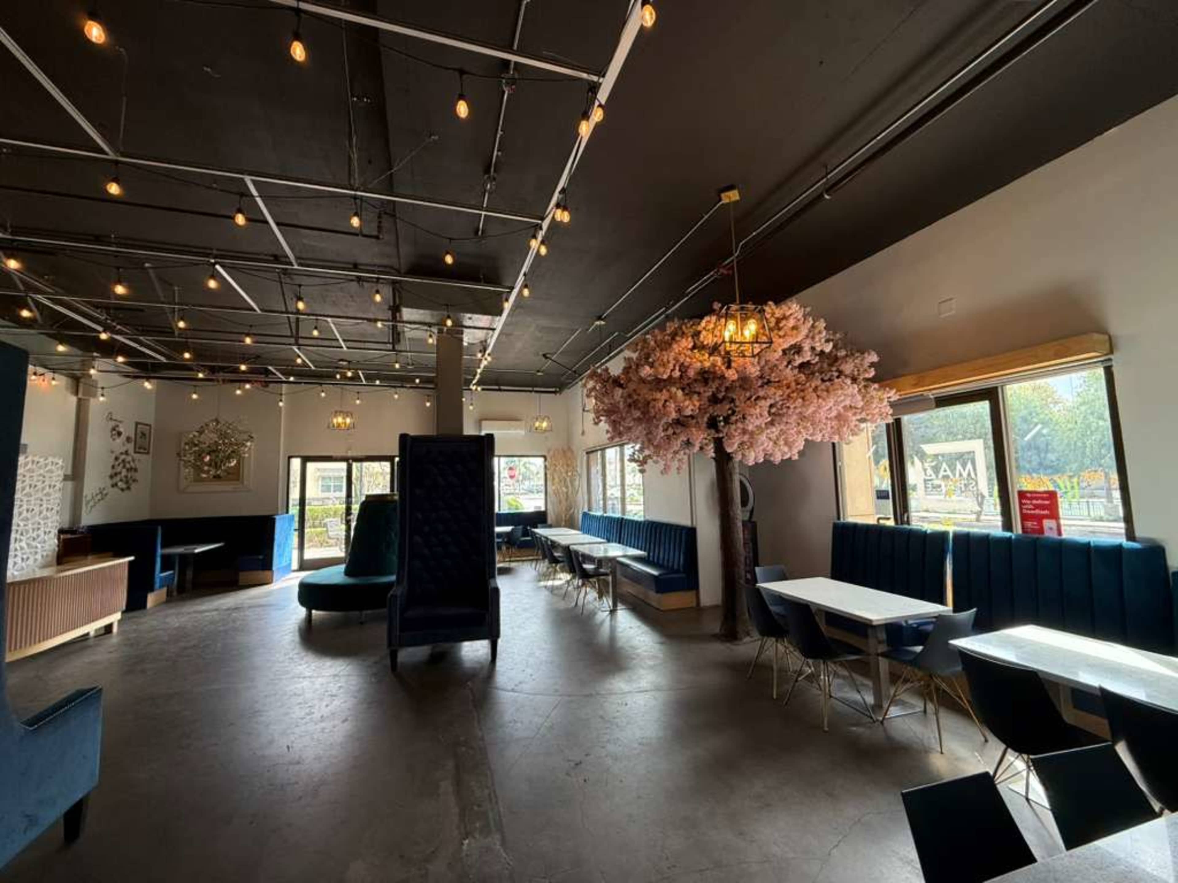 The image shows a spacious, modern café interior with blue seating, a large pink cherry blossom tree, and decorative lighting.