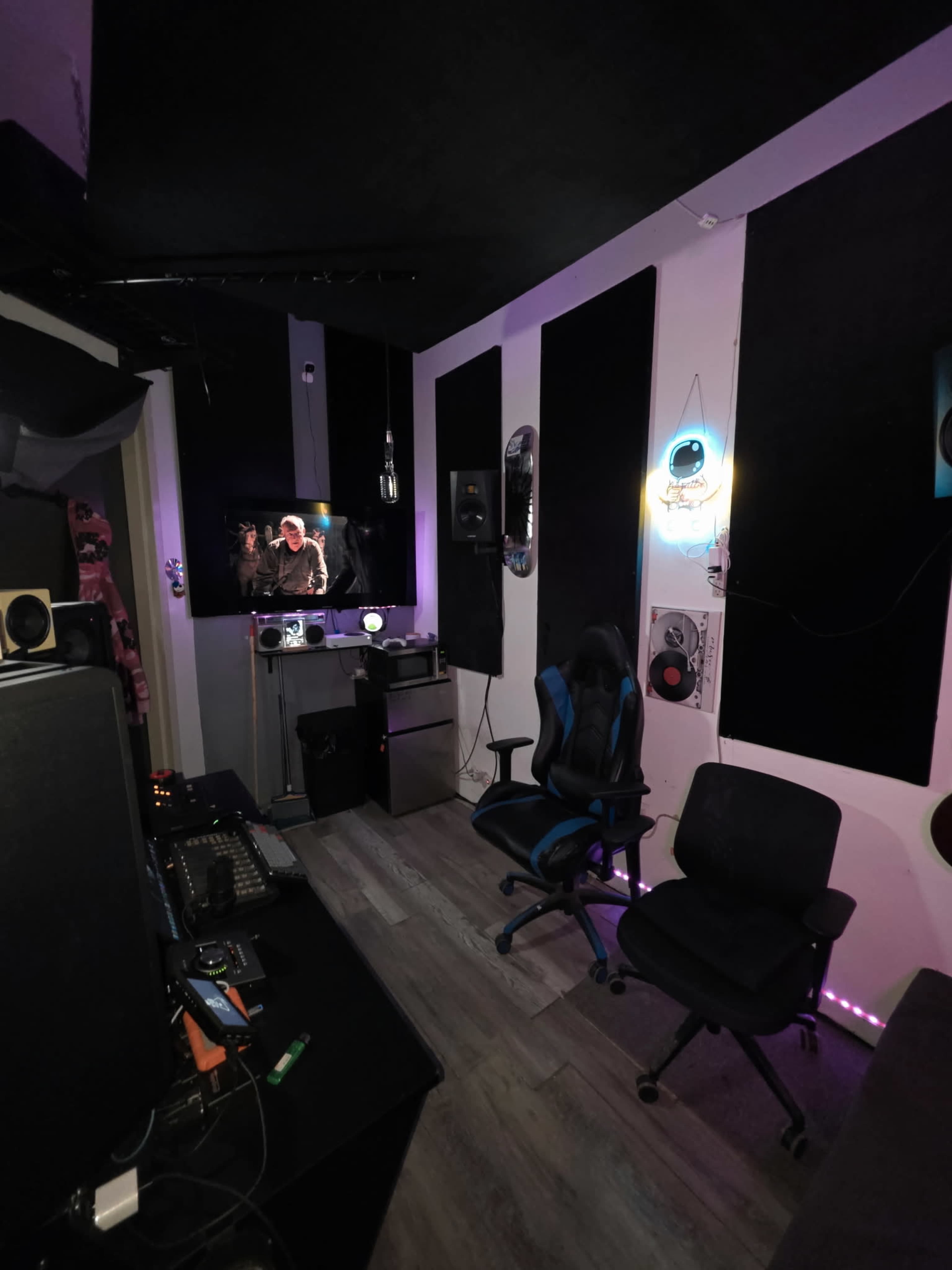 The image shows a dimly lit room with soundproof panels on the walls, a television displaying a scene, and two chairs near a desk with audio equipment.