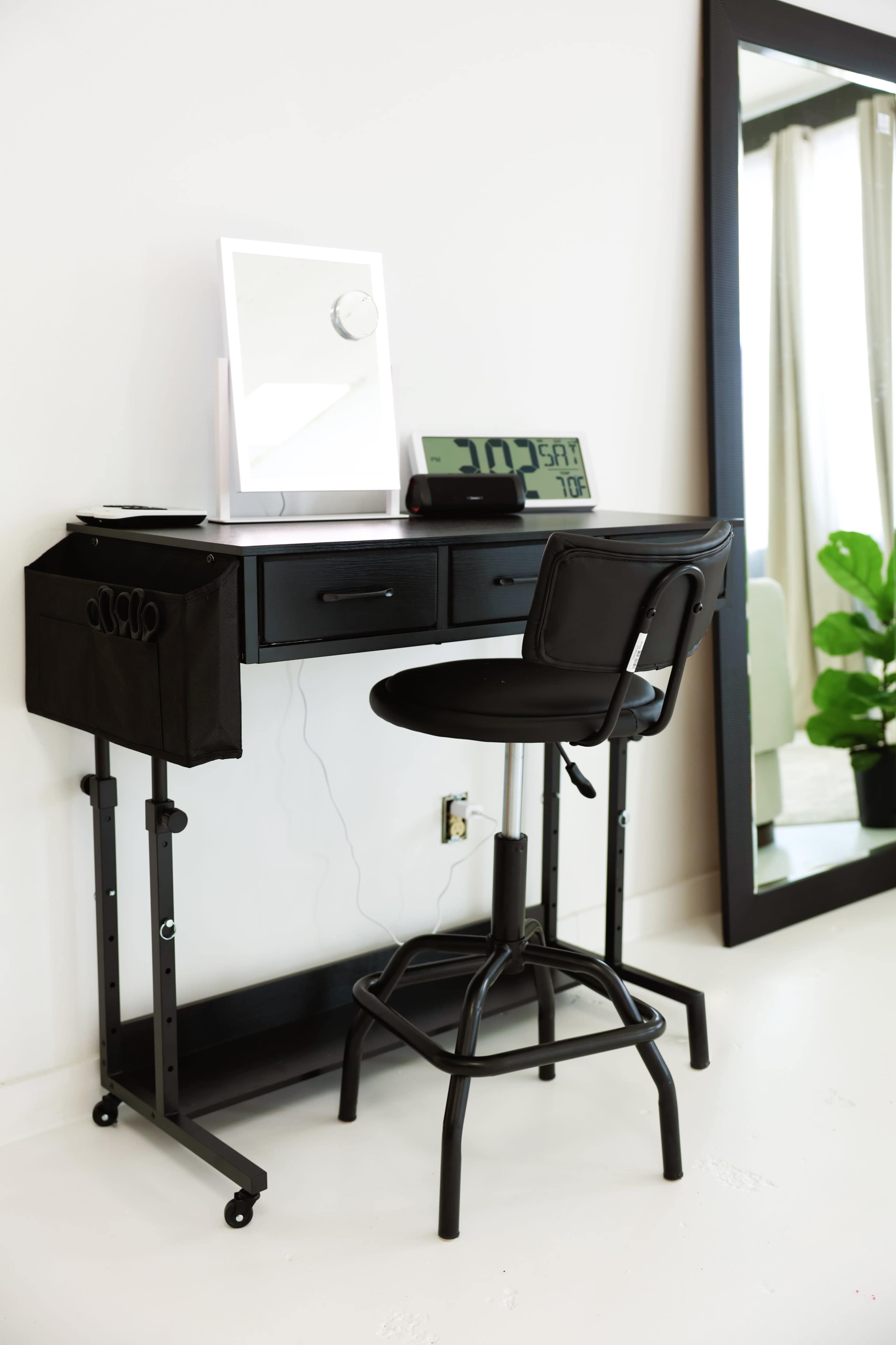 The image shows a compact black desk with a mirror and lamp on top, accompanied by a swivel stool and a large mirror against a white wall, while a green plant is visible nearby.