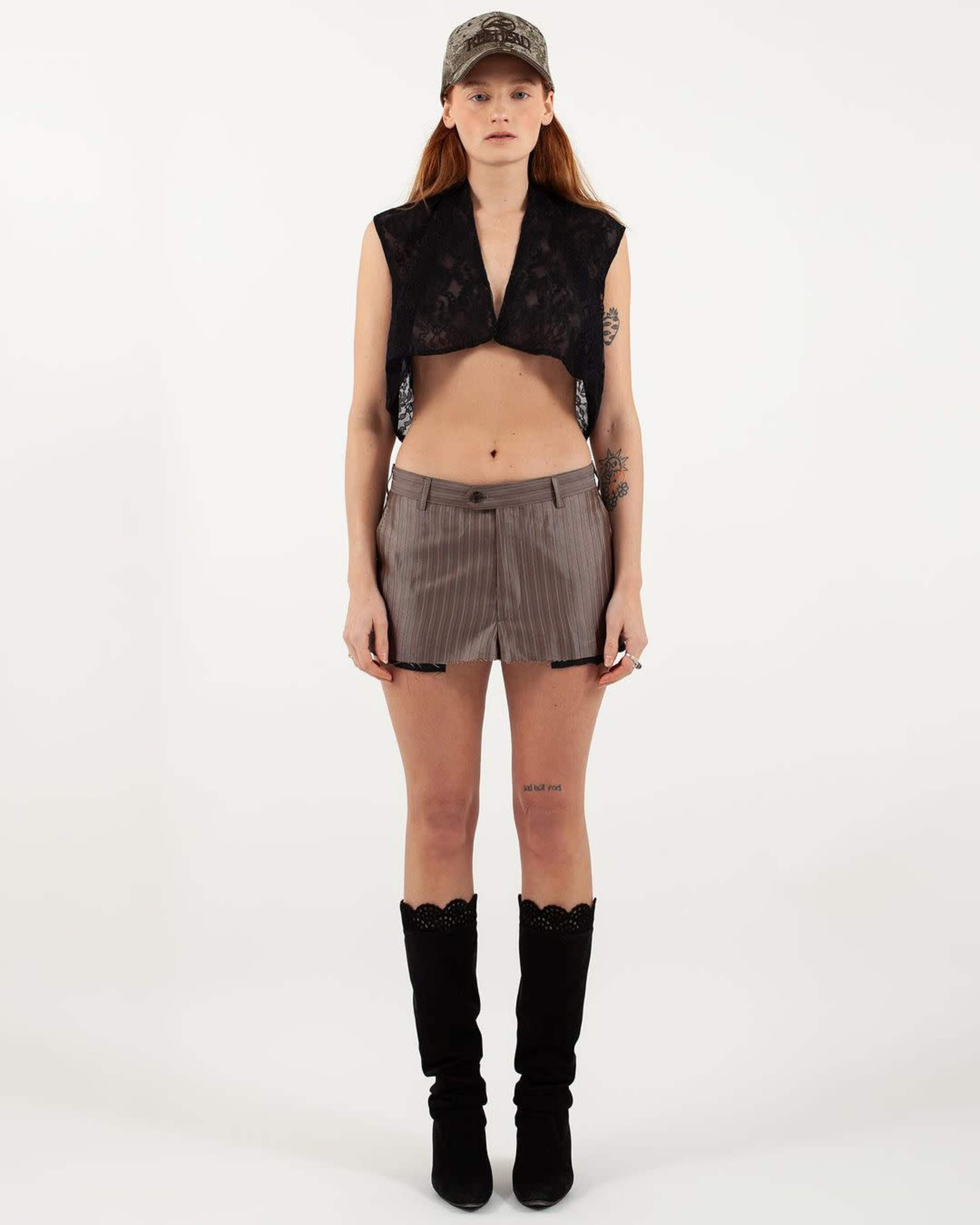 A model stands upright wearing a cropped black lace top, striped shorts, a camouflage cap, and knee-high black boots.