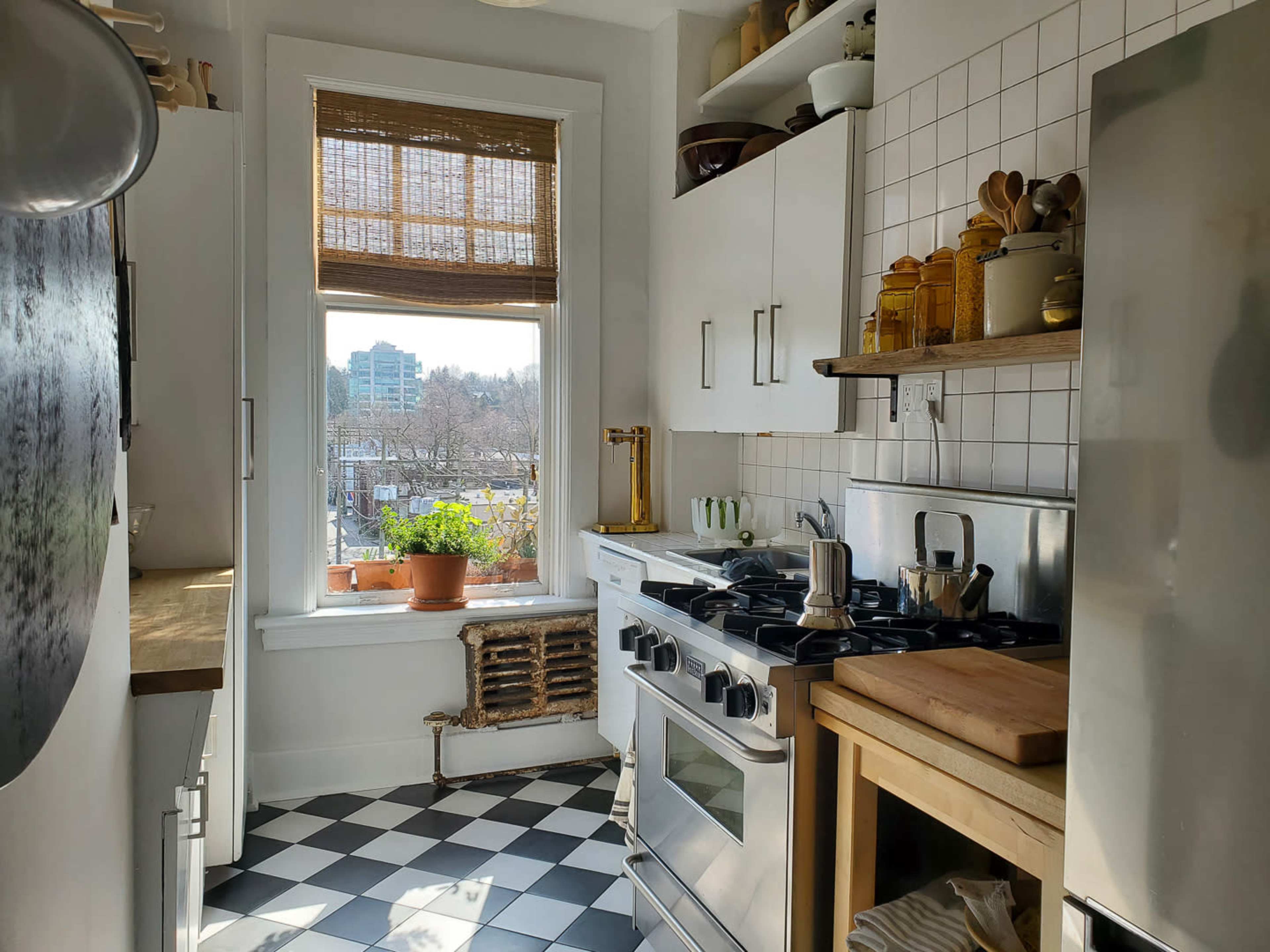 A narrow kitchen features a stove, wooden countertop, and a window with a view of the city, illuminated by natural light.