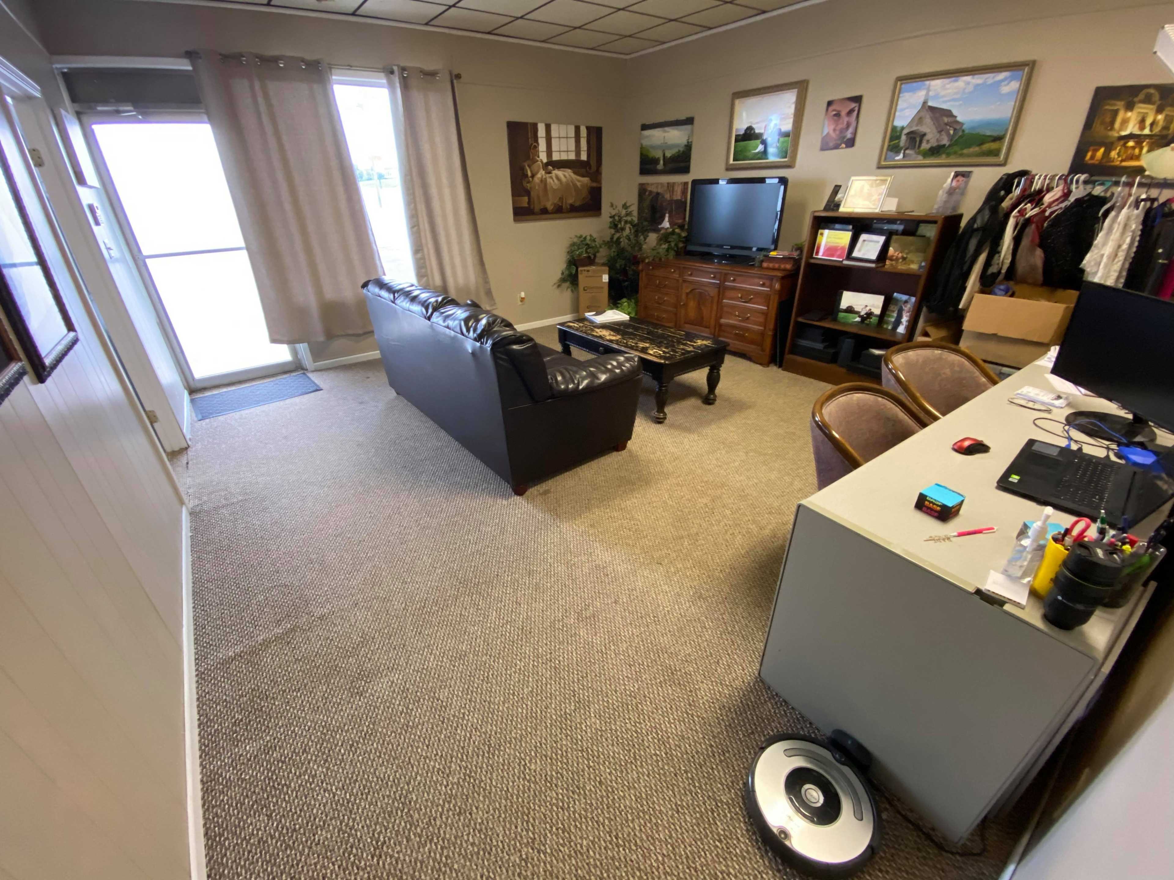 The image shows an office space featuring a black leather sofa, a wooden dresser, a television, a desk with a computer, and a doorway leading outside.