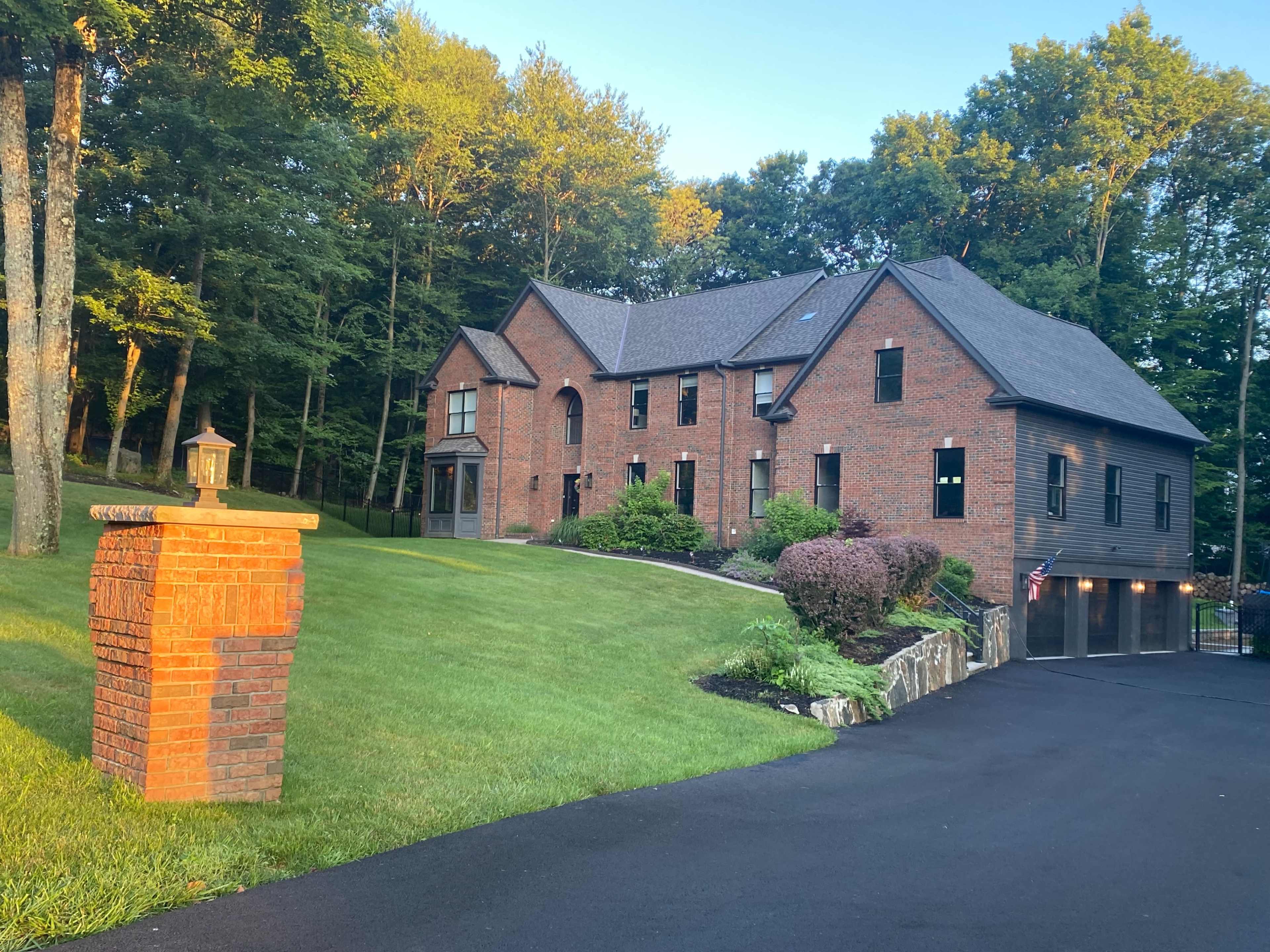 A two-story brick house with landscaped gardens sits on a green lawn next to a paved driveway.