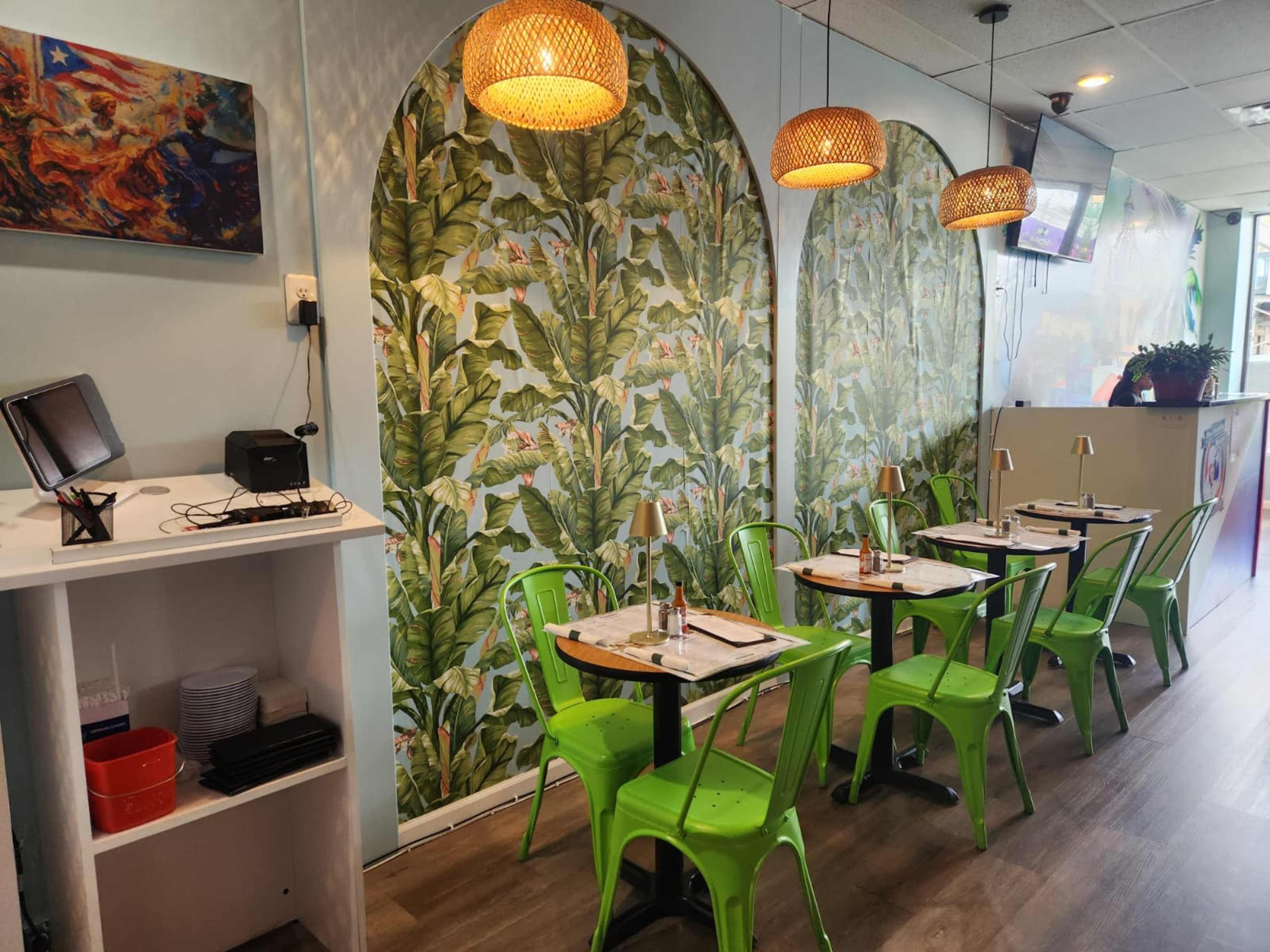 The image shows a dining area with green metal chairs and tables, set against a wall featuring tropical leaf wallpaper and decorative pendant lights.