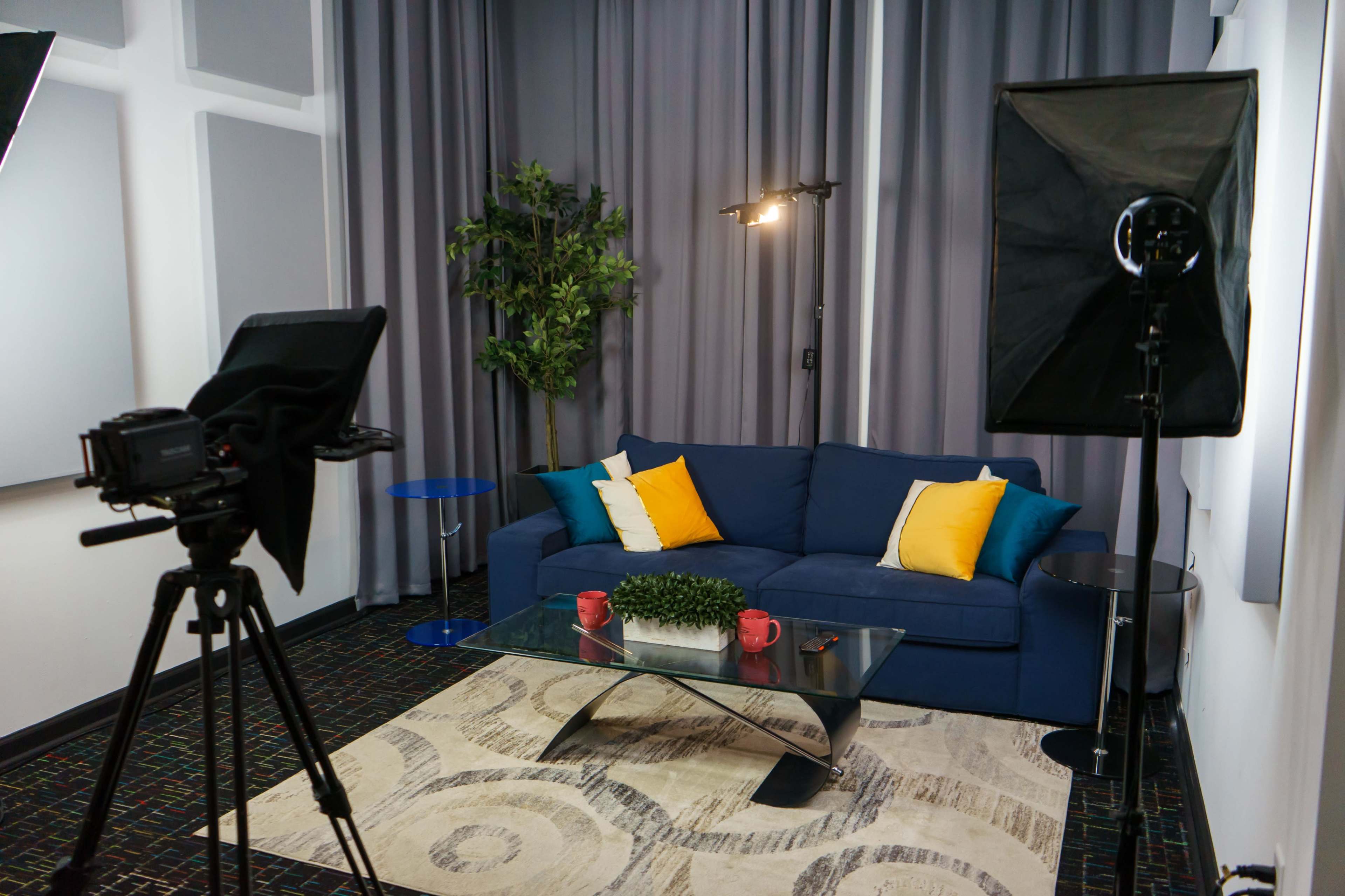 A modern interior setup with a blue sofa adorned with yellow cushions, a glass coffee table, and studio lights positioned around a small plant in a corner.