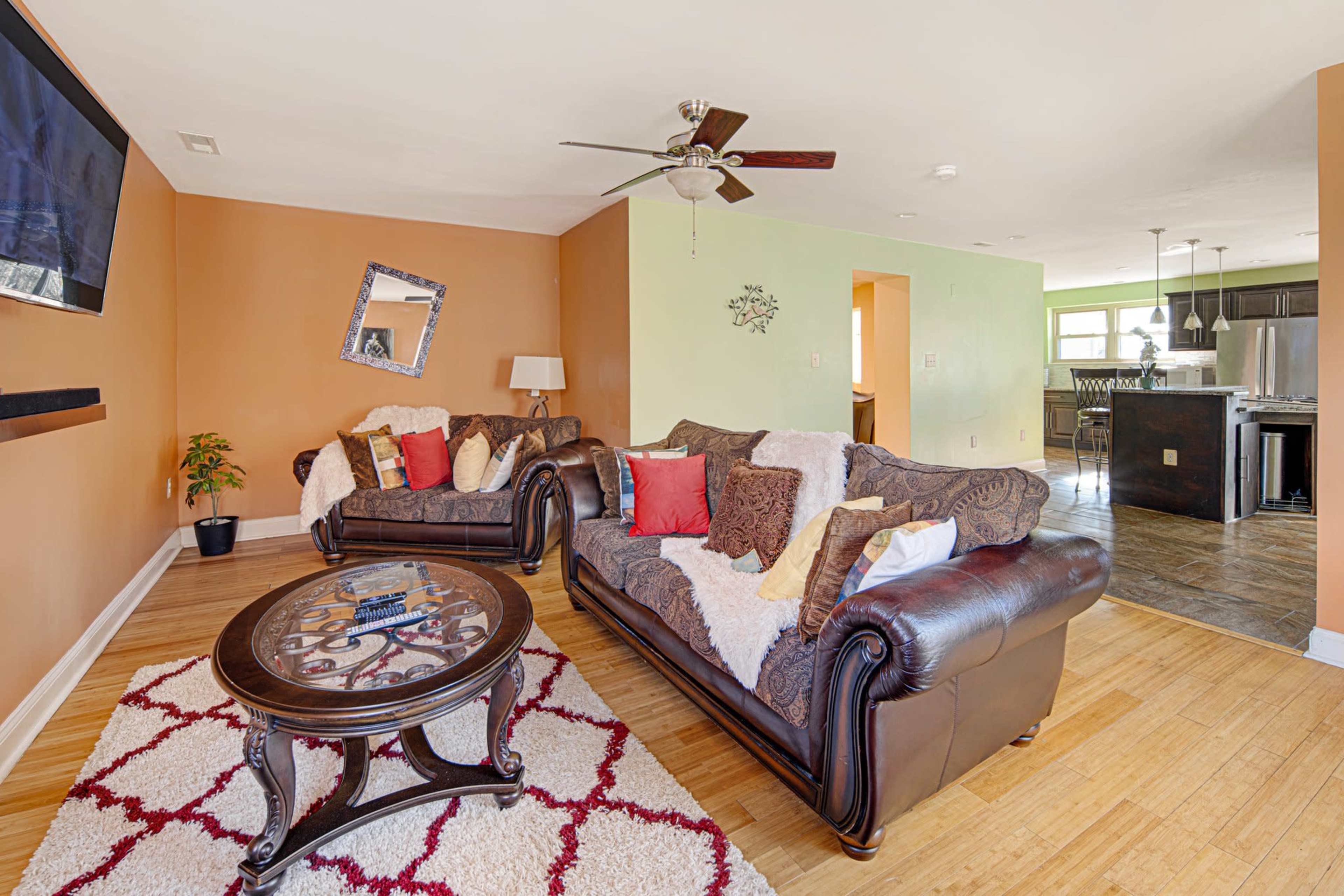 The living room features two dark leather sofas with decorative pillows arranged around a round coffee table on a patterned rug, with an open kitchen visible in the background.