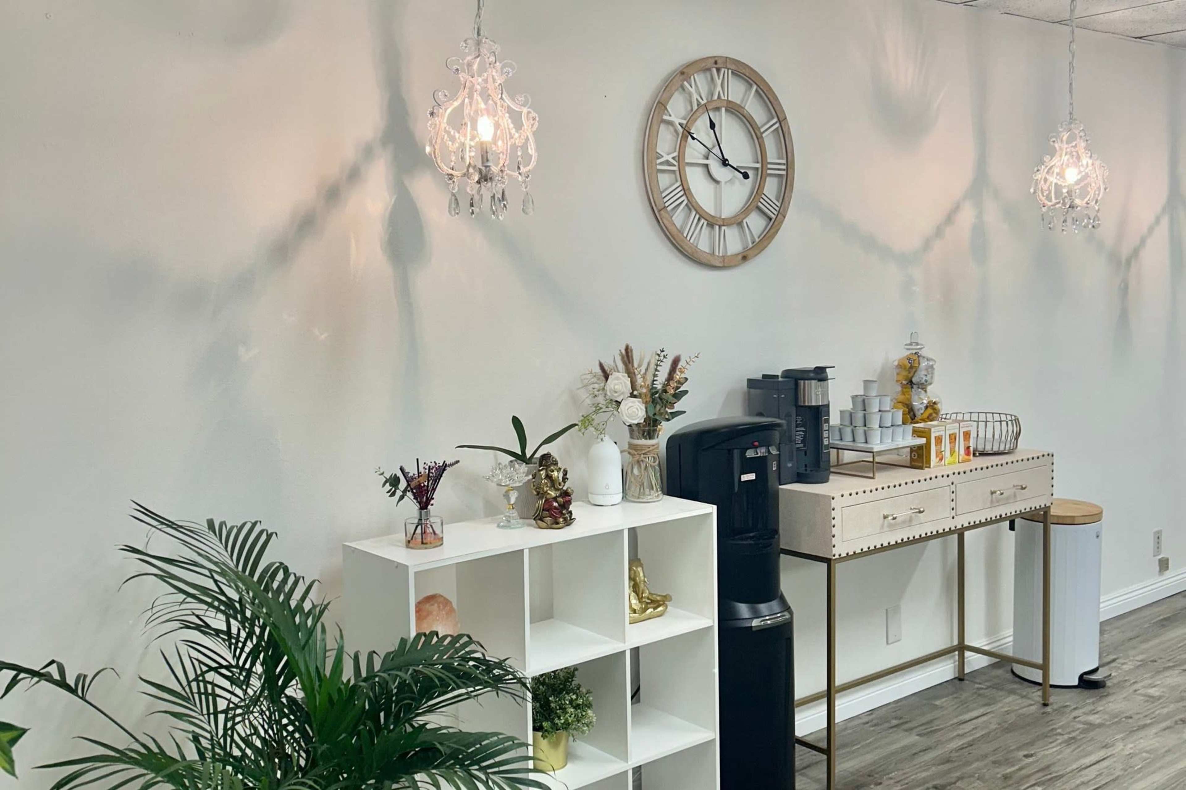 The image shows a well-decorated room featuring a wall clock, decorative lighting fixtures, a coffee station, and a white shelving unit with plants and ornaments.
