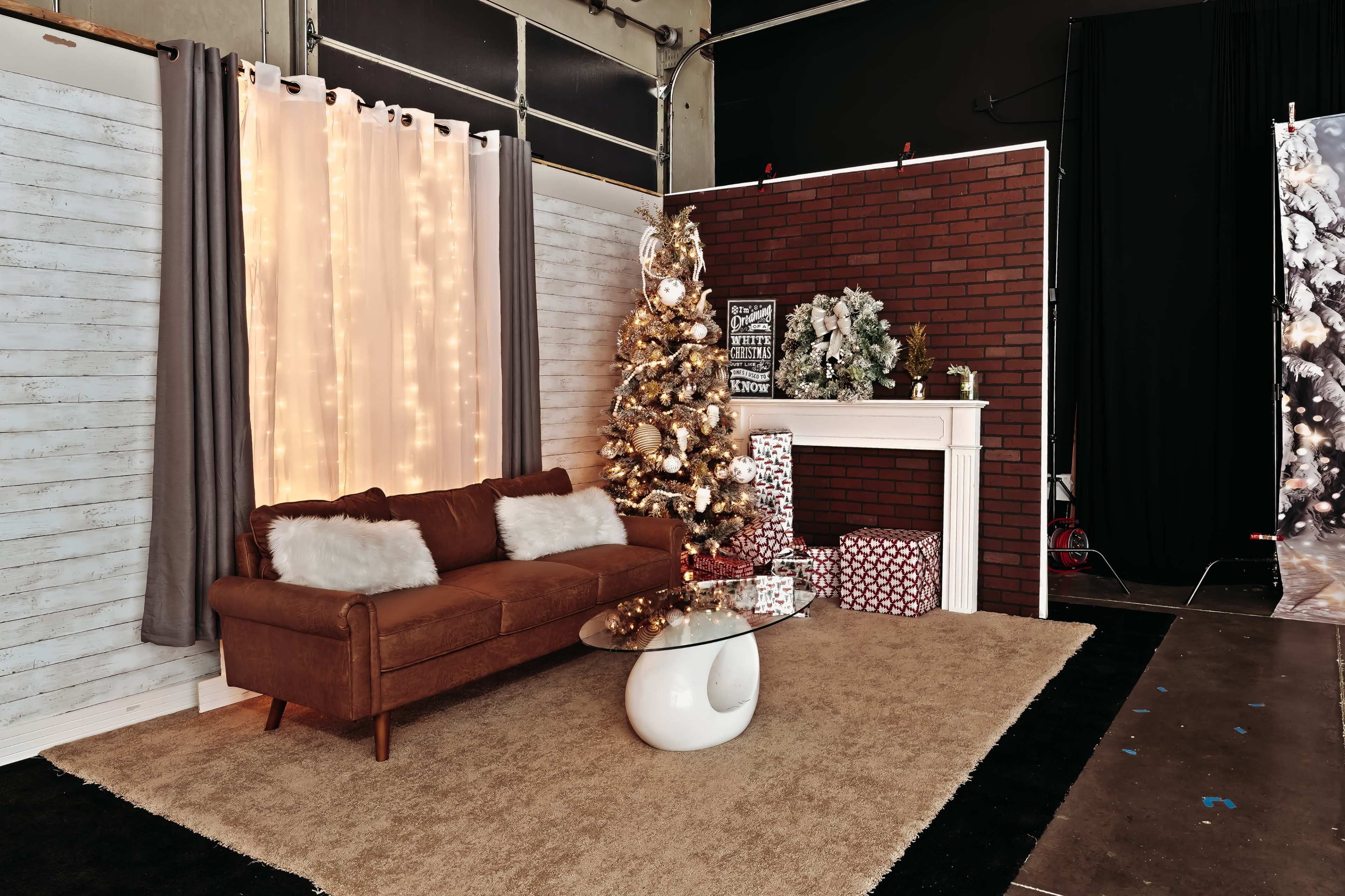 A cozy room setup featuring a brown sofa, a glass coffee table, a decorated Christmas tree, and a white fireplace with presents underneath.
