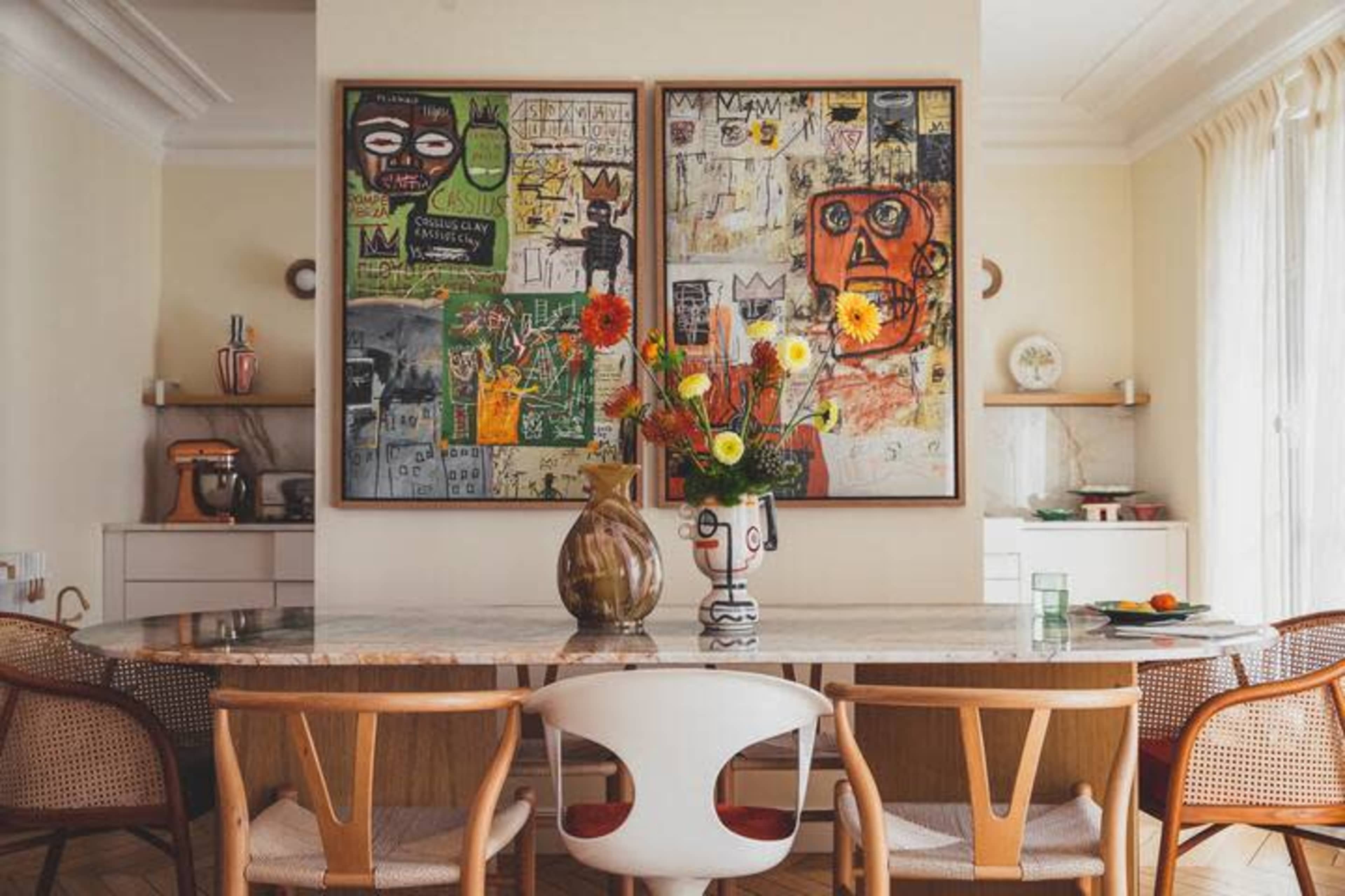 A modern dining area features a large table surrounded by wooden chairs, with colorful artwork on the walls and a floral centerpiece.