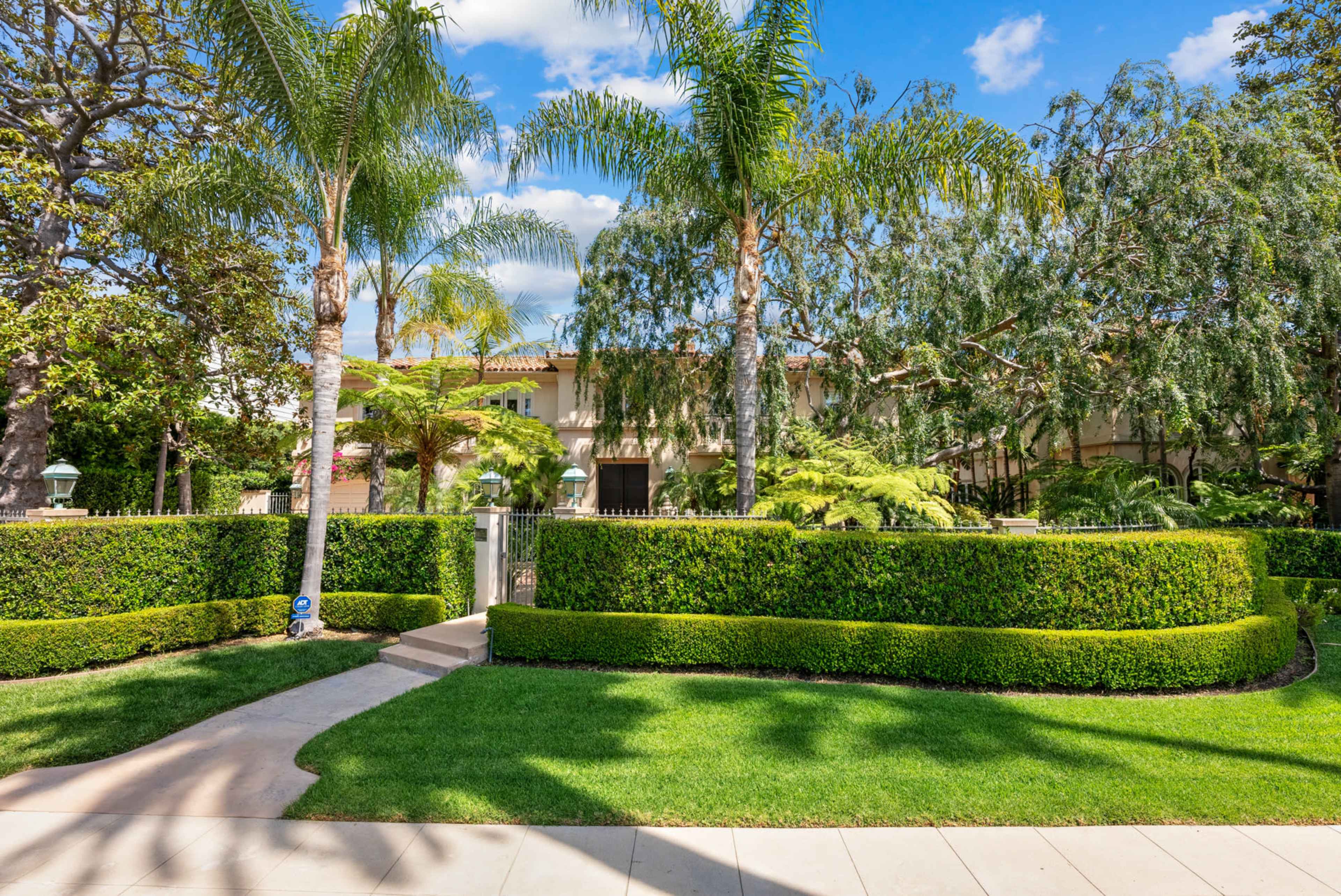 The image shows a landscaped front yard with neatly trimmed hedges, palm trees, and a pathway leading to a large house partially obscured by greenery.