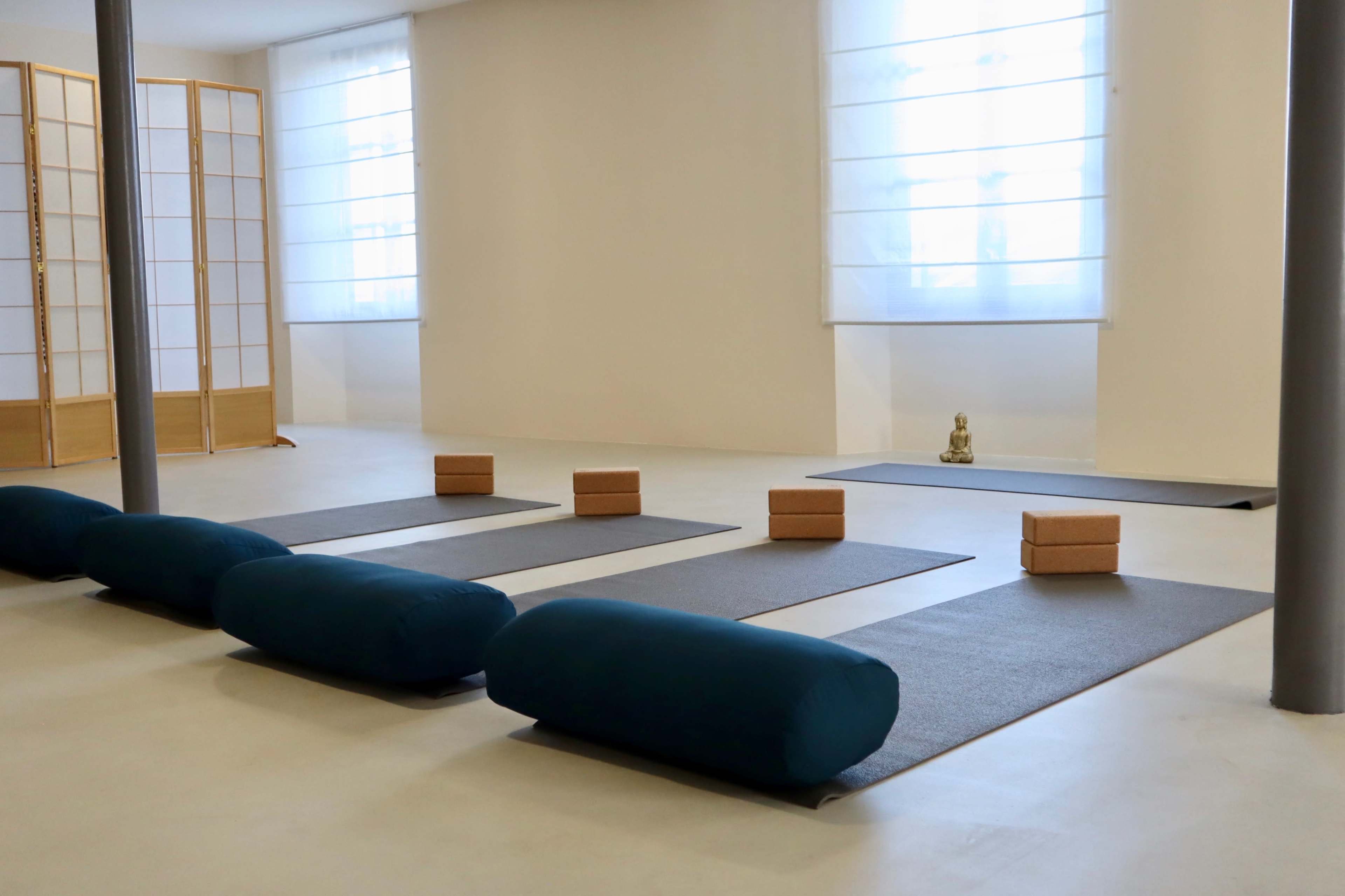 A tranquil yoga or meditation studio with blue bolsters, gray mats, and blocks arranged around a small Buddha statue.