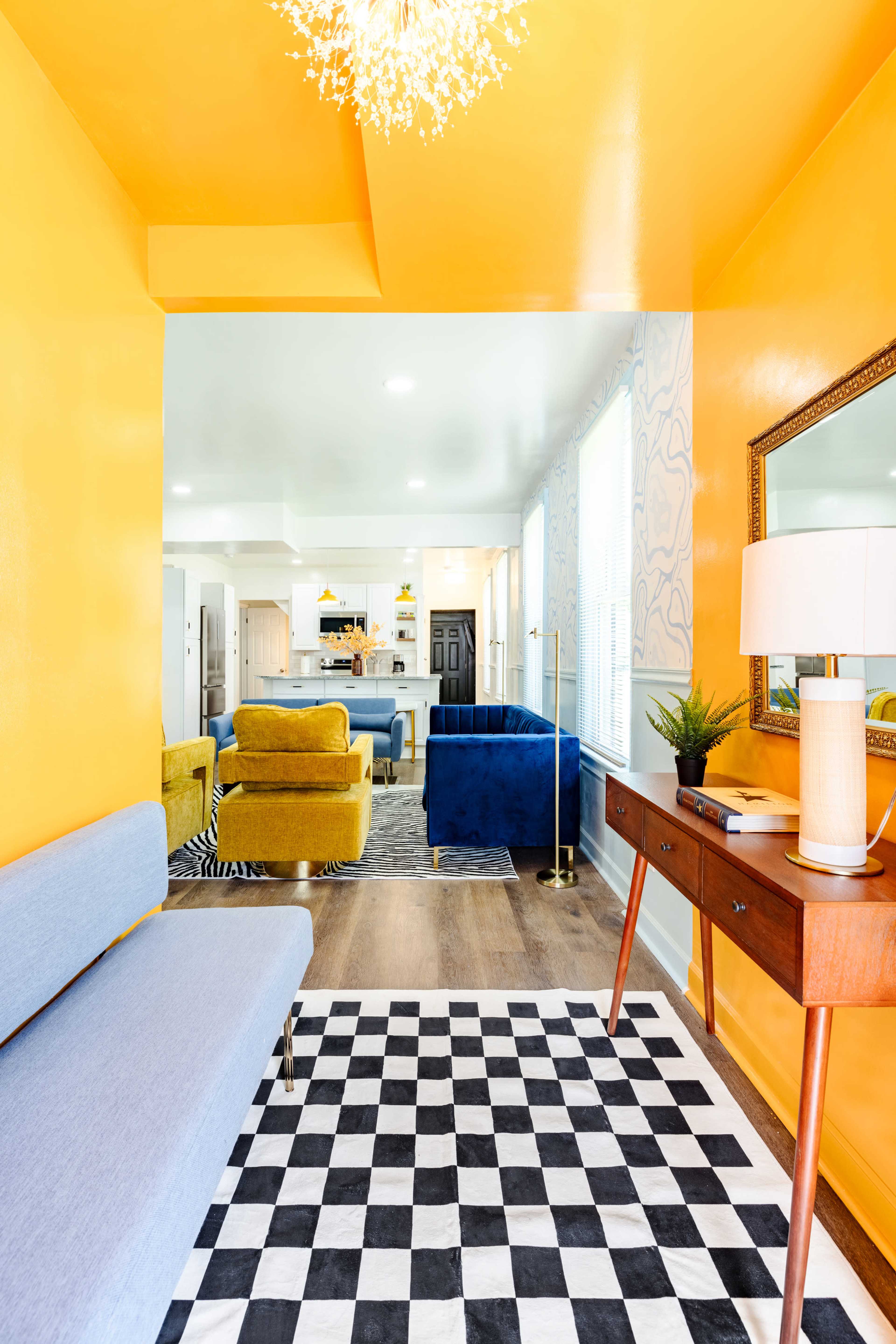 A brightly colored room with yellow walls, featuring a blue sofa and a black-and-white checkered rug, leading to a living area with additional colorful furniture.