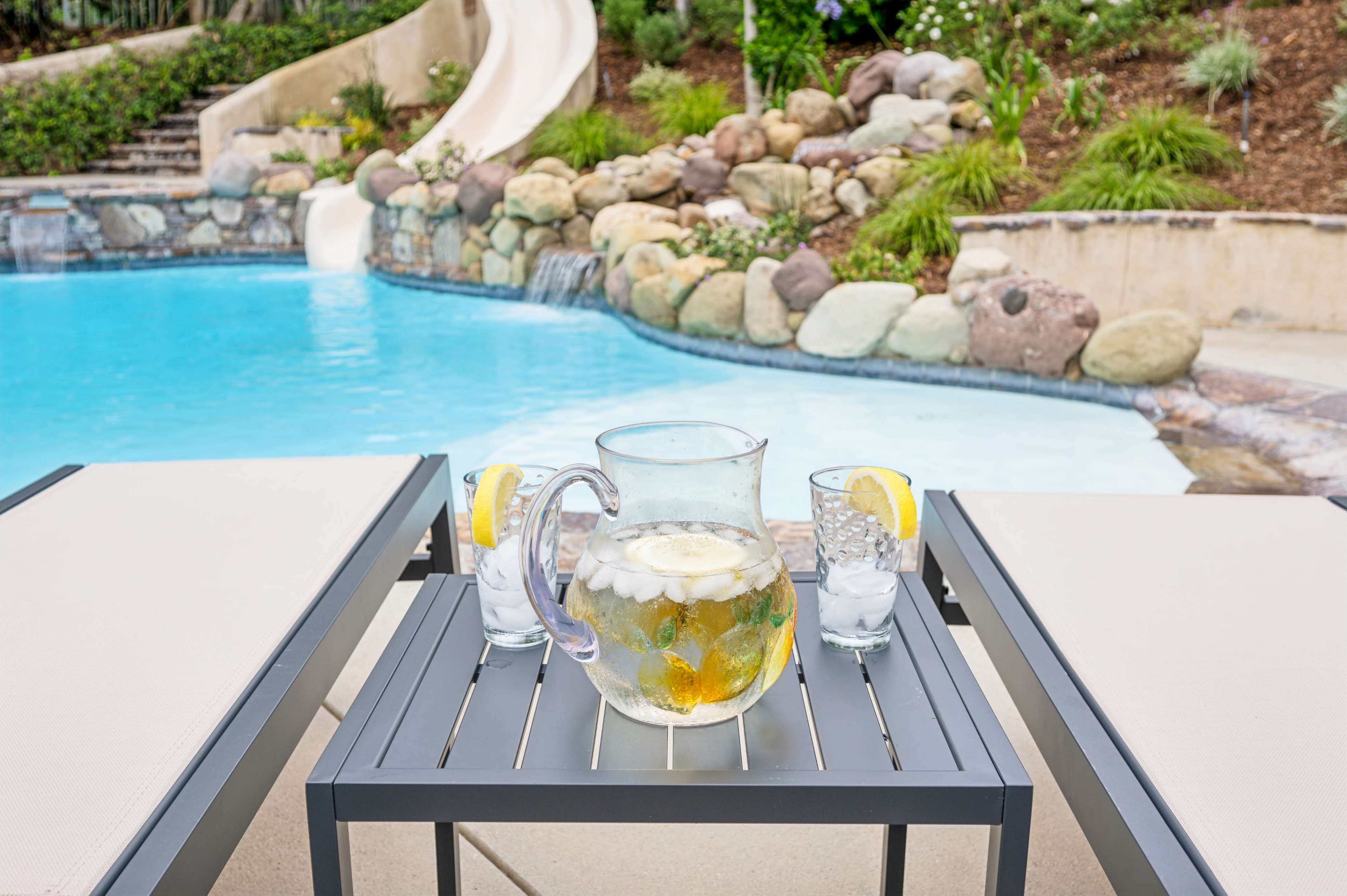 A pitcher of lemonade with lemon slices and ice sits on a small black table next to two glasses, beside a blue swimming pool and stone slide.