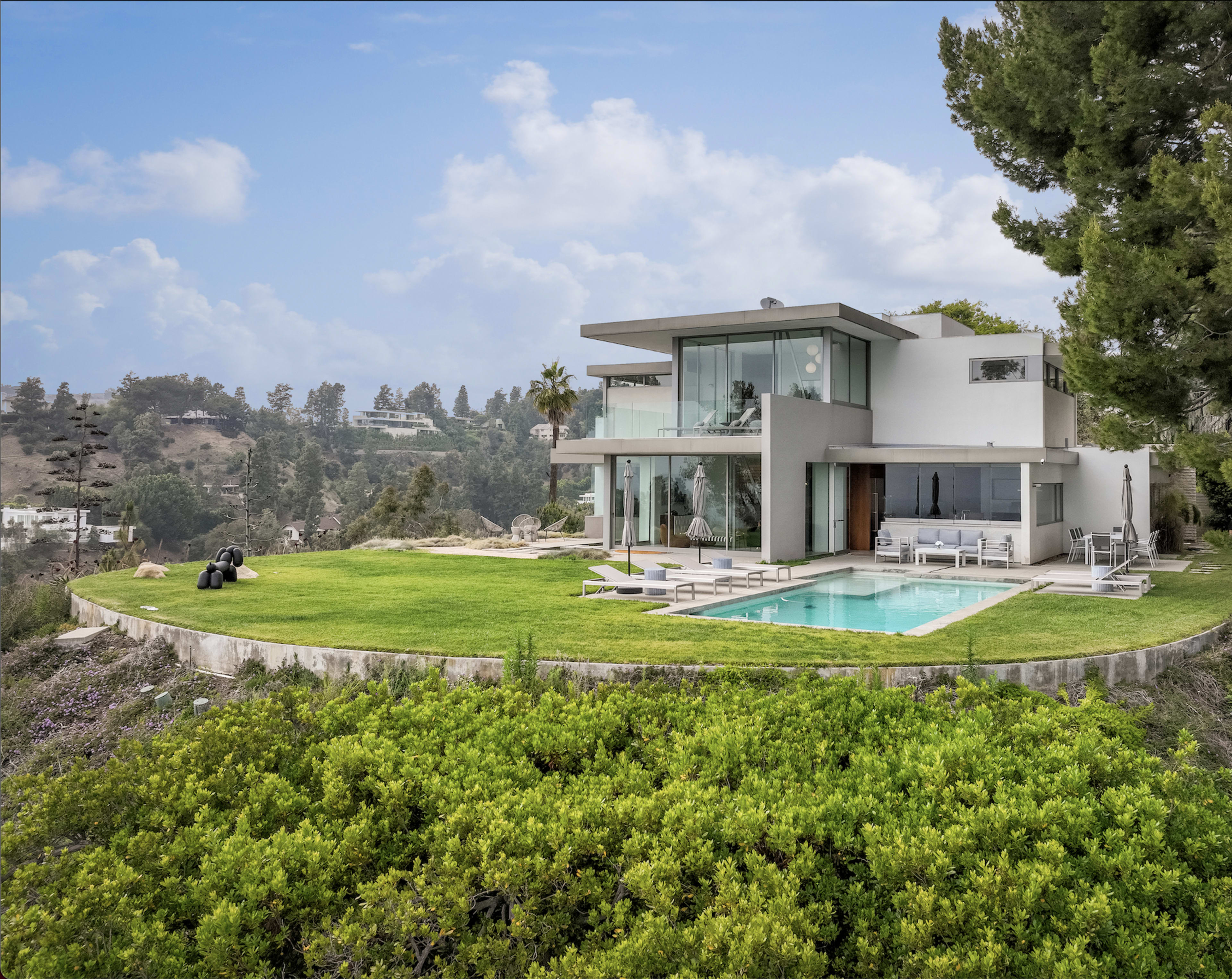 A modern house with large windows and a swimming pool is situated on a hillside, surrounded by greenery.