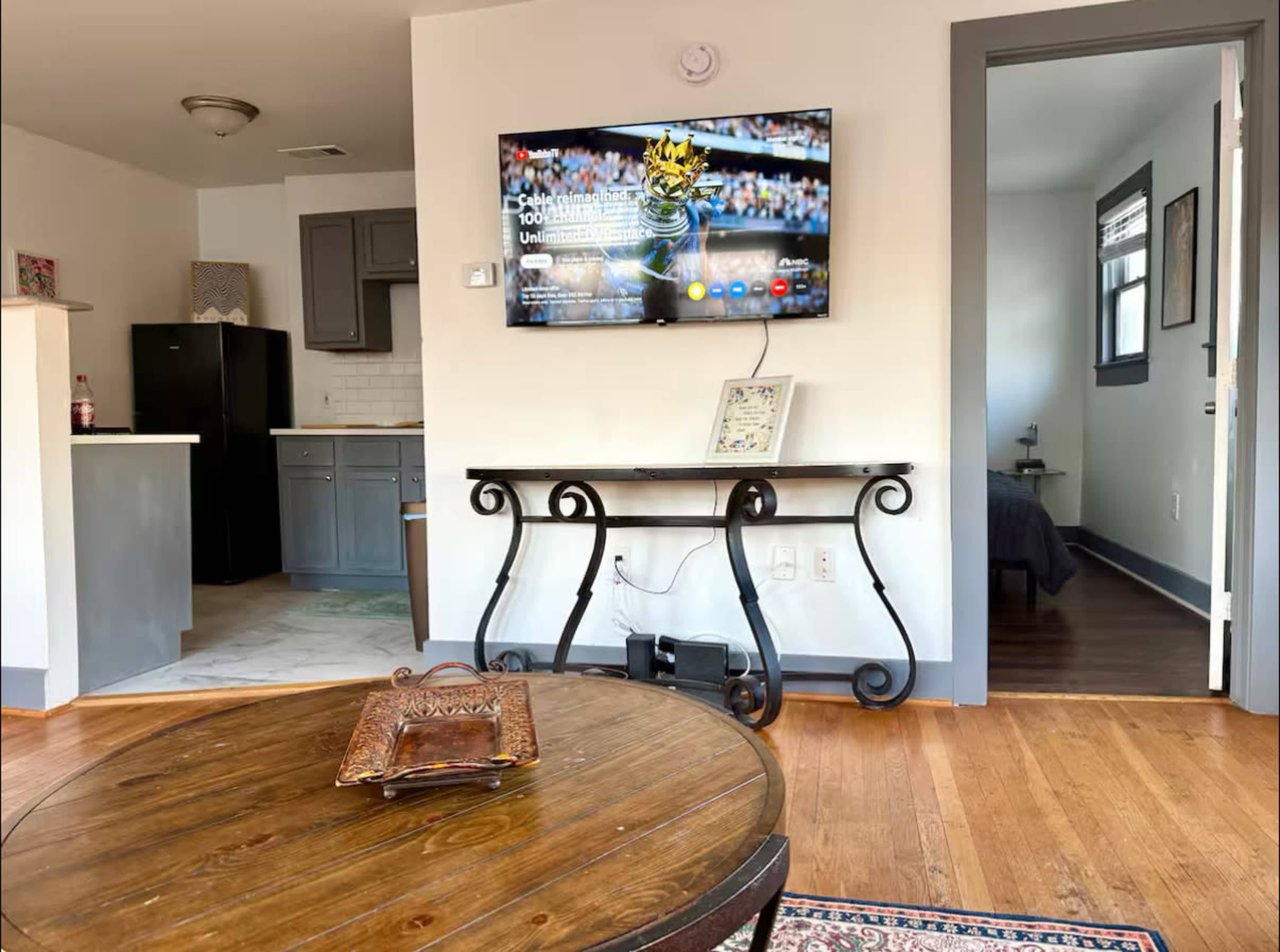 The image shows a living room with a wall-mounted TV displaying content, a wooden coffee table in the foreground, and an open doorway leading to another room.