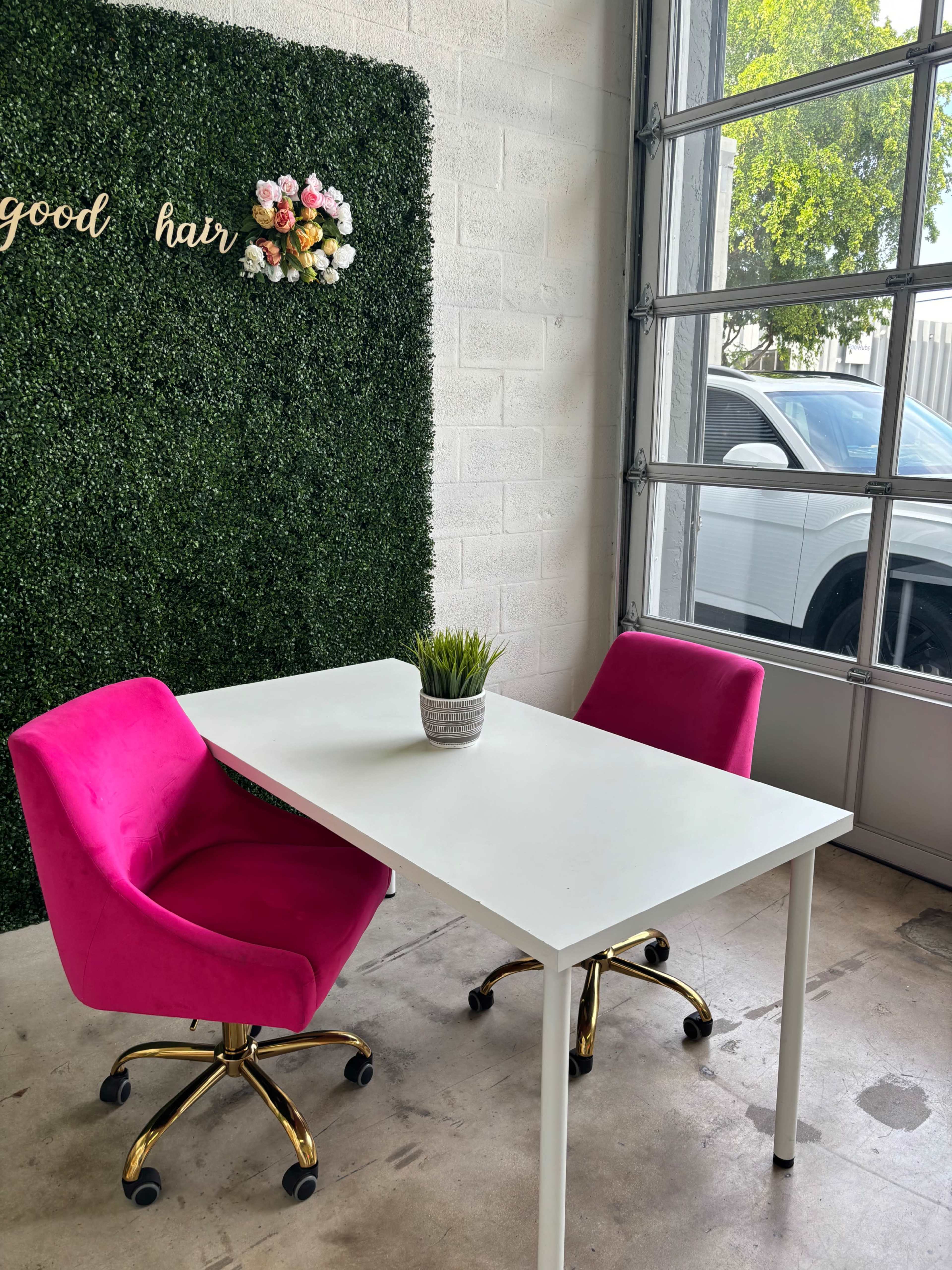 A minimalist workspace features a white table with two pink chairs, a potted plant, and a green wall adorned with floral accents.